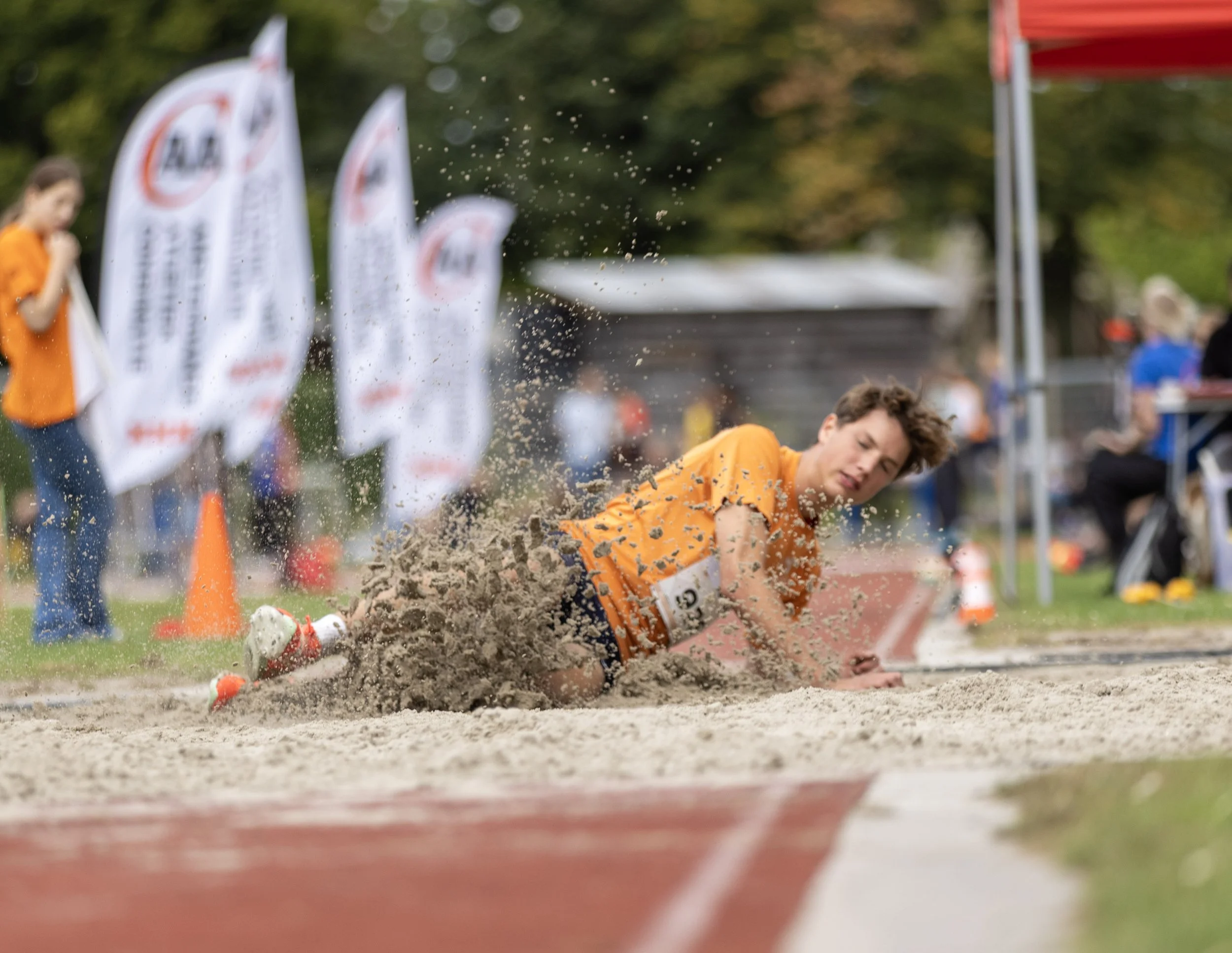 A young male athlete falling and landing in a sandpit during a long jump event at an outdoor track and field competition, with spectators and officials in the background.