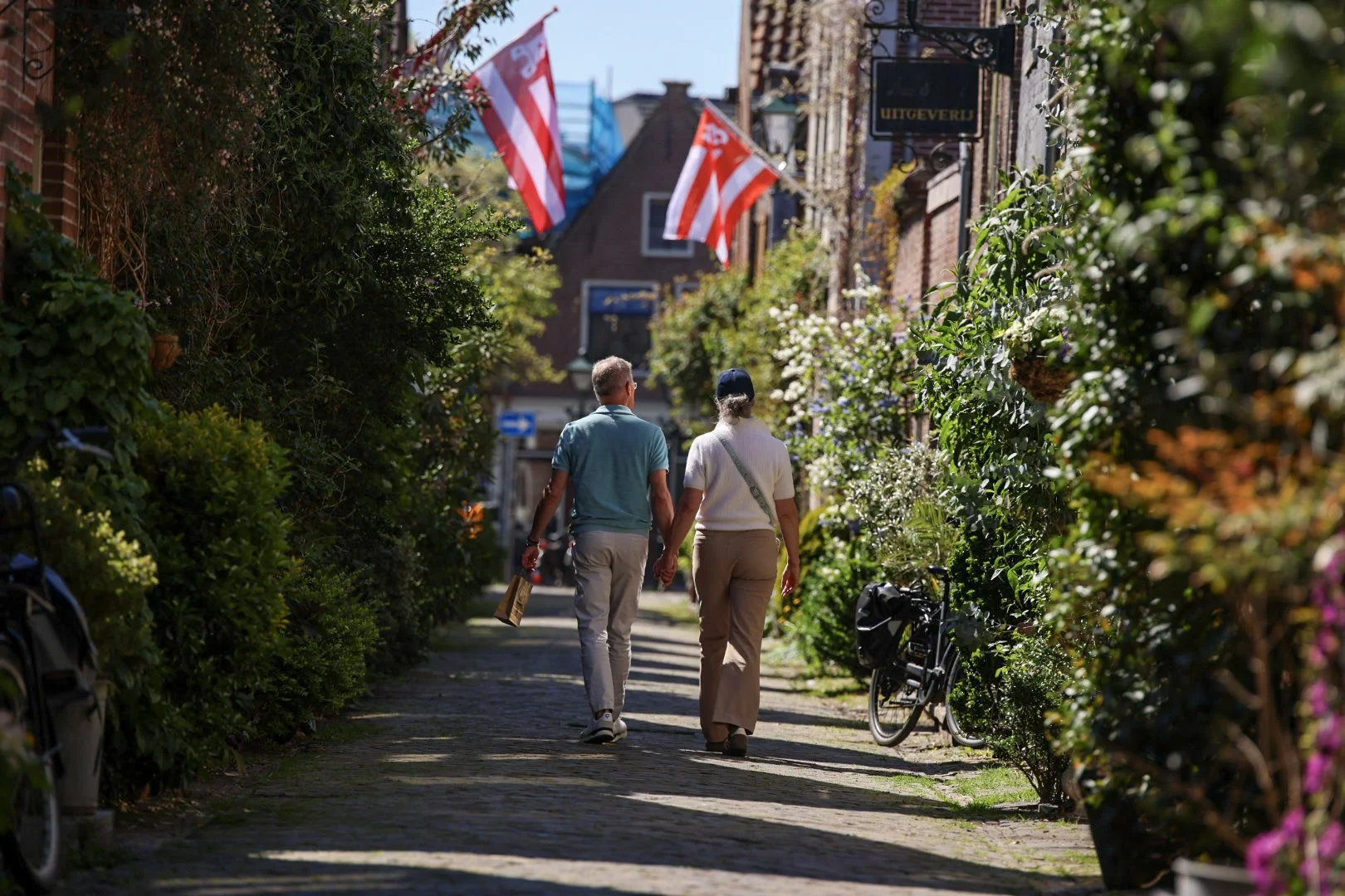 A couple walking down a charming, tree-lined street with lush greenery and bushes on both sides, holding hands. The street features flags hanging above, a bicycle parked on the side, and brick buildings with signs, creating a cozy, scenic atmosphere.