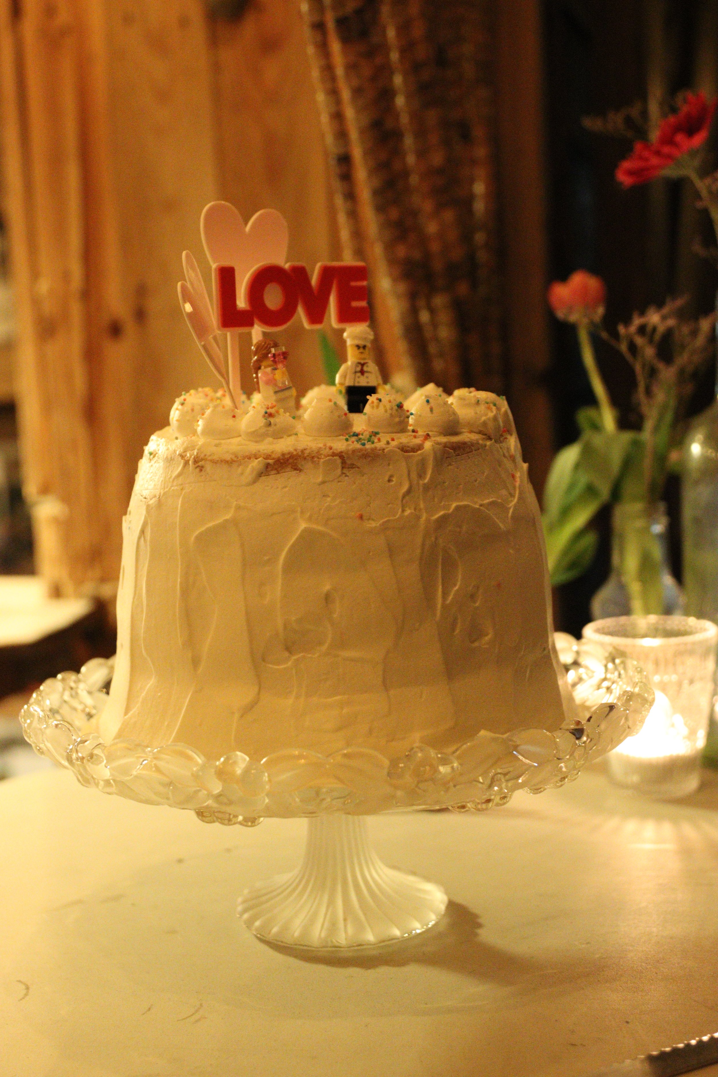 A wedding cake decorated with small doll figures, including a bride and groom, and a sign that reads 'LOVE'. The cake has white icing with sprinkles and is displayed on a white ornate cake stand.