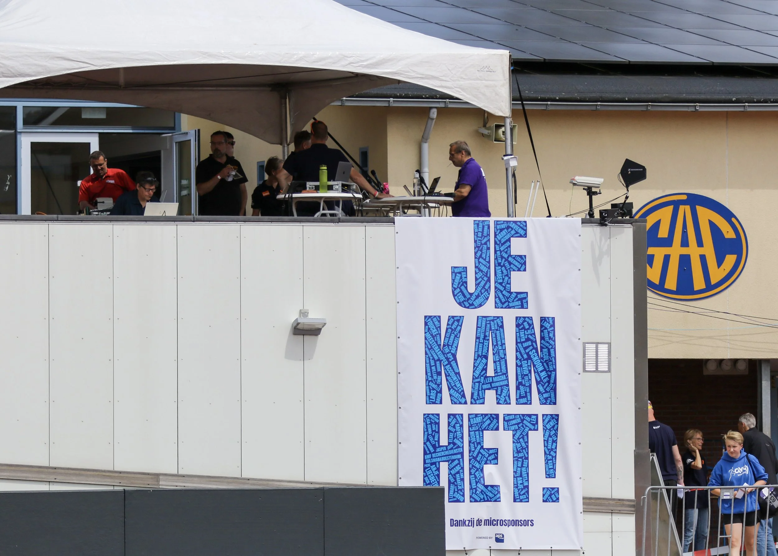 People gathered on a rooftop terrace with DJ equipment, and a large white banner with blue text saying 'JEG KAN IKKE!' hung on the wall, with some individuals visible in the background.