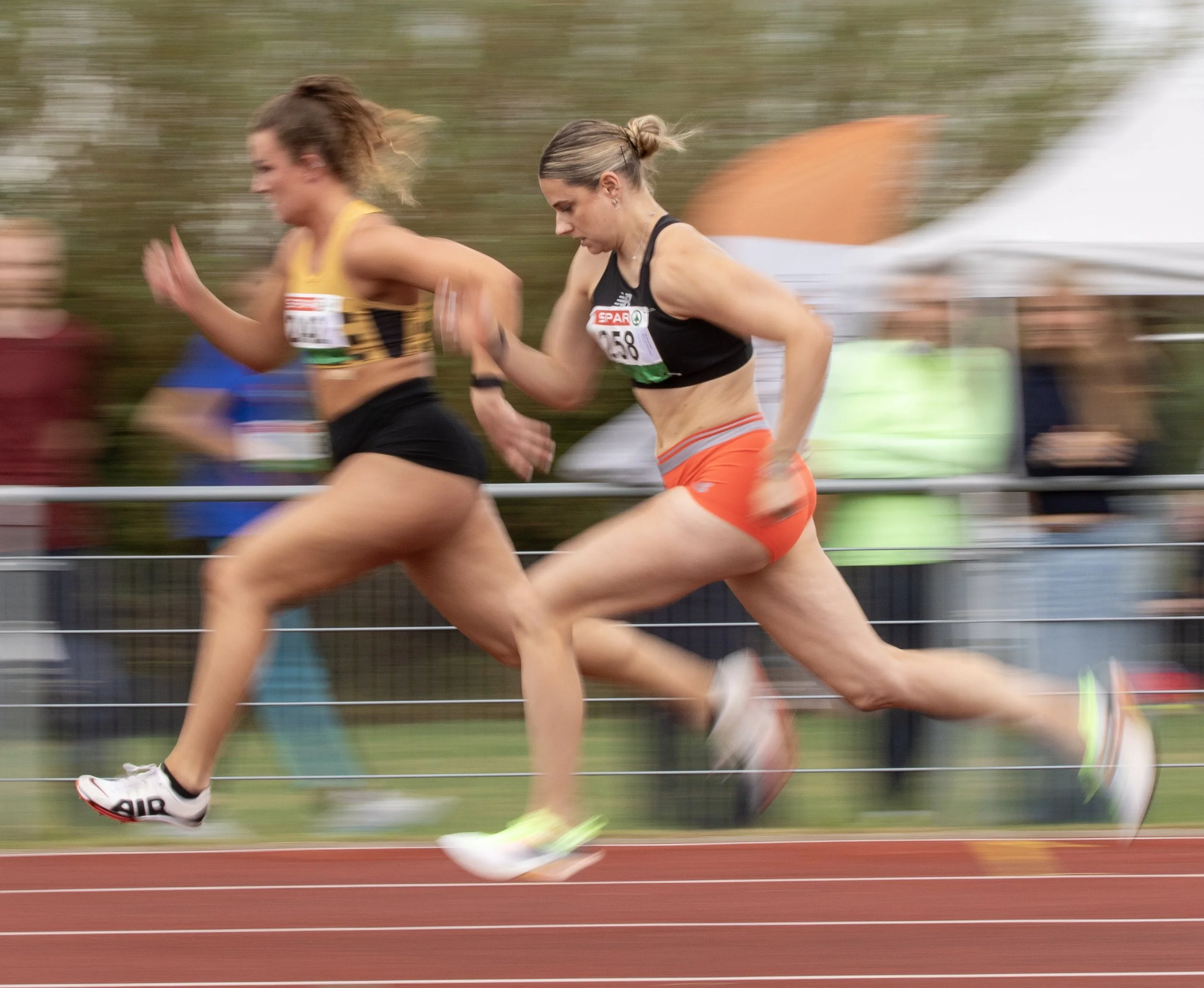 Two female athletes competing in a track race, running side by side at full speed on a red track with a white barrier, with spectators and tents in the background.