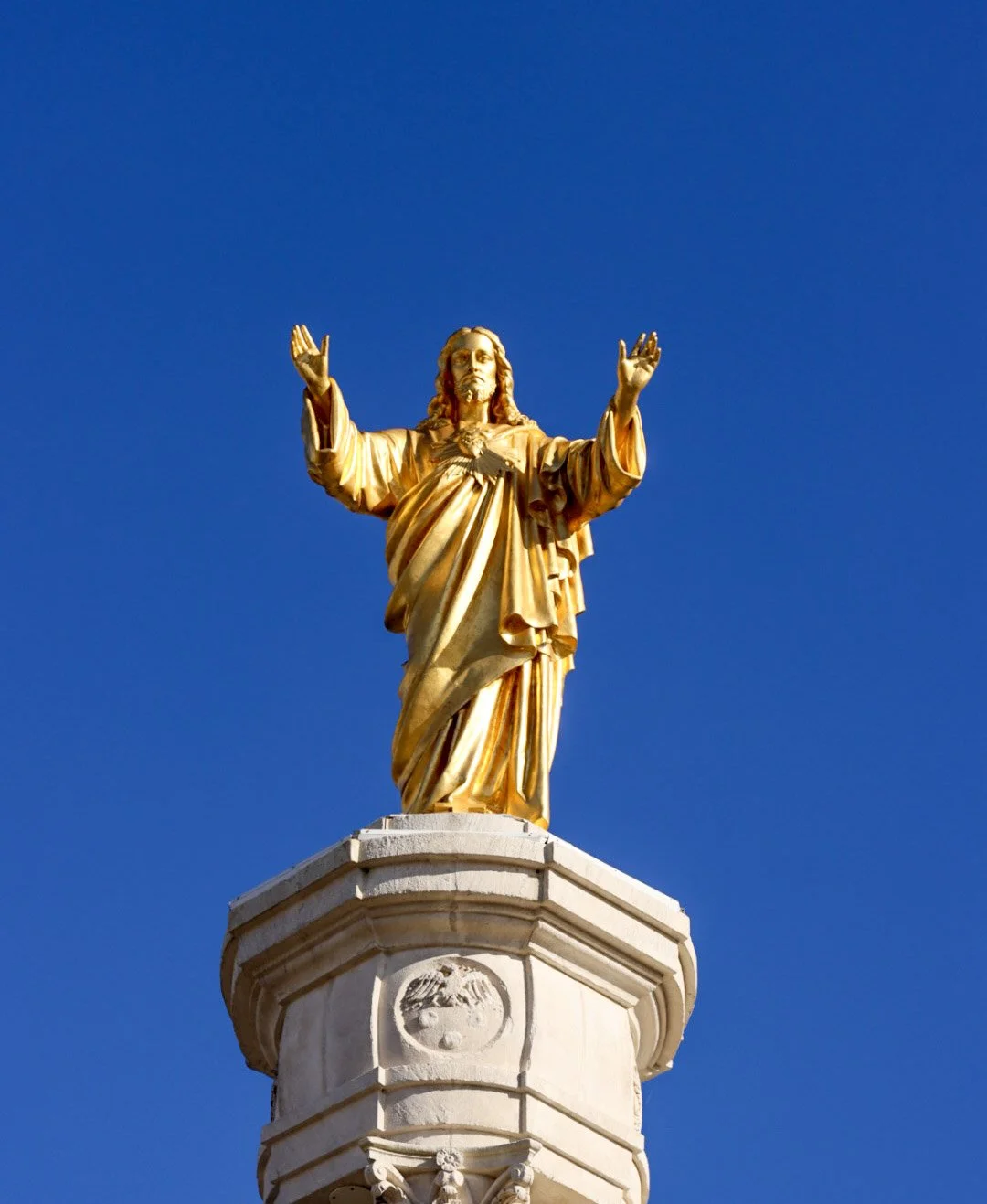 Golden statue of Jesus Christ with open arms on a white pedestal against a clear blue sky.