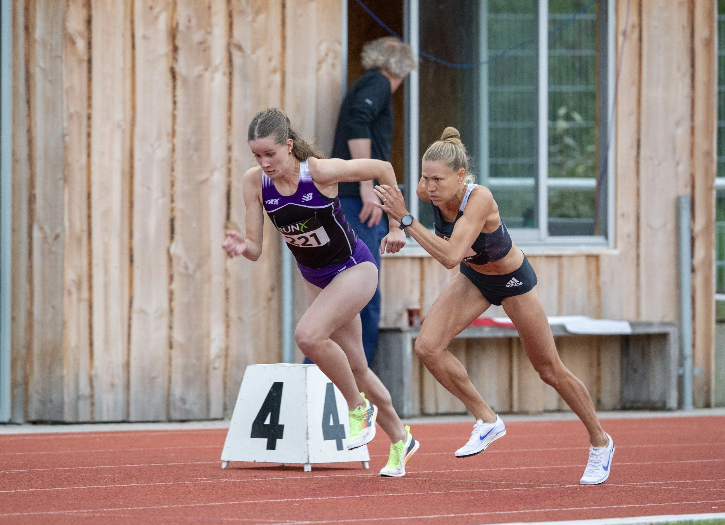 Two female athletes competing in a sprint race on an indoor track, with a coach or official in the background.