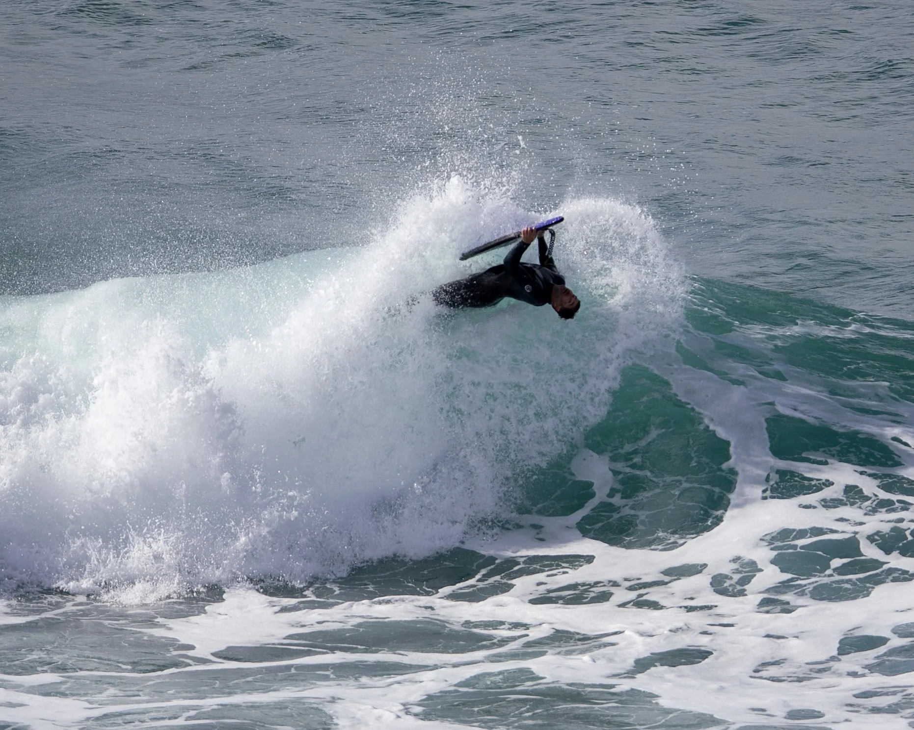 A person wearing a wetsuit is performing a flip on a surfboard in the ocean, surrounded by splashing water.