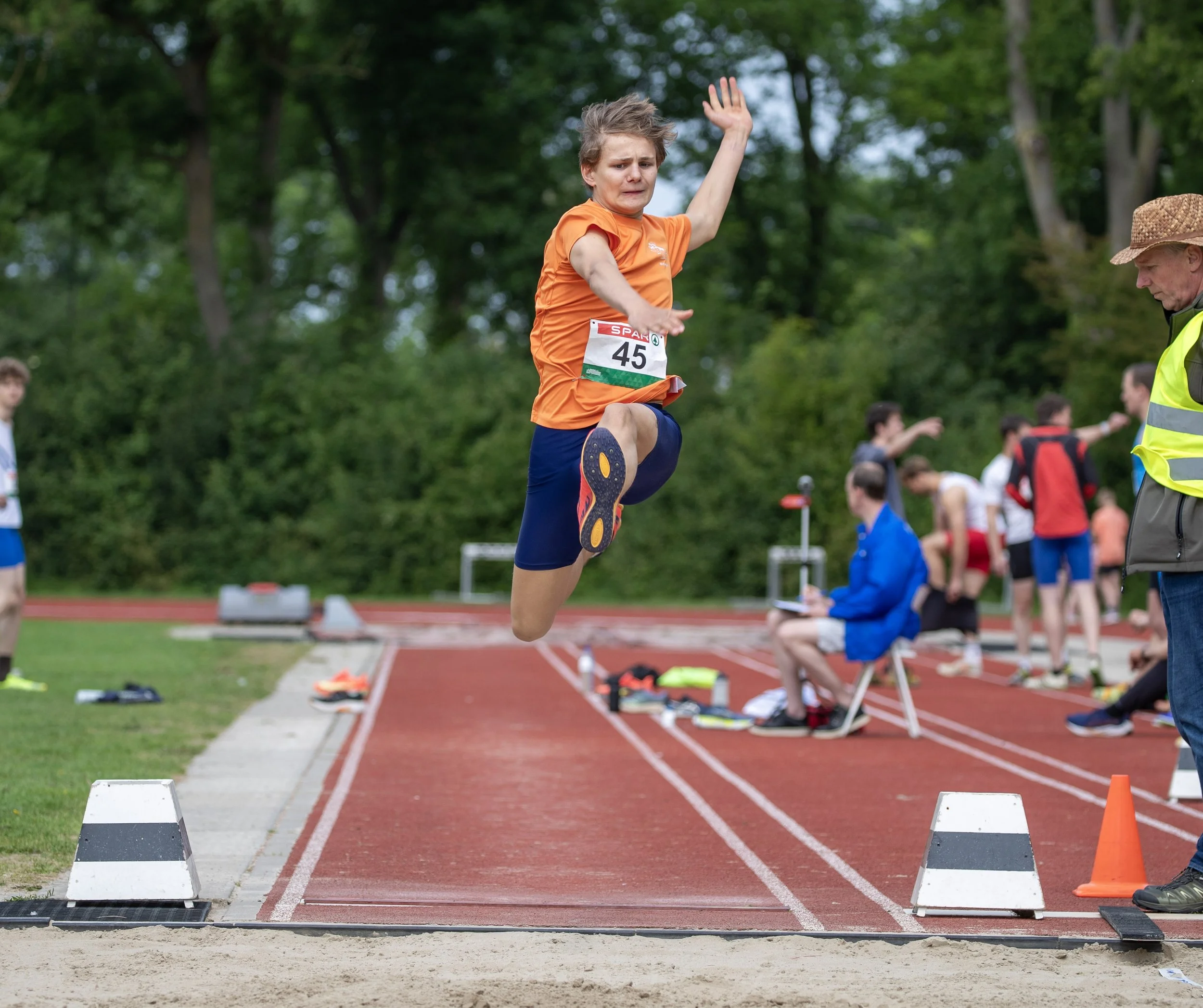 A male athlete in an orange shirt and blue shorts is mid-air during a long jump at a track and field event, with a group of spectators and other competitors in the background.