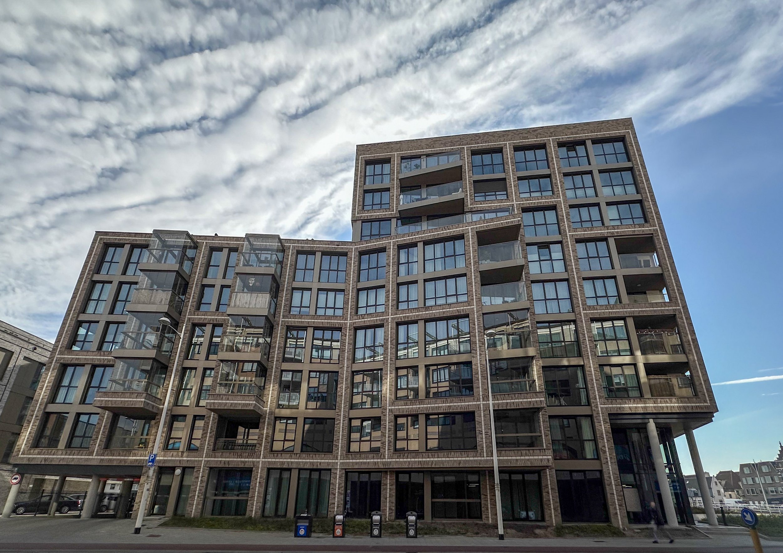 Modern multi-story apartment building with large glass windows and balconies under a partly cloudy sky.