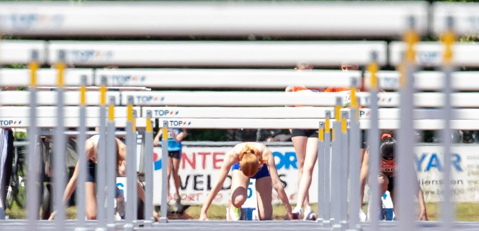 Track athletes preparing at the starting line for a race, viewed through hurdles on a track.