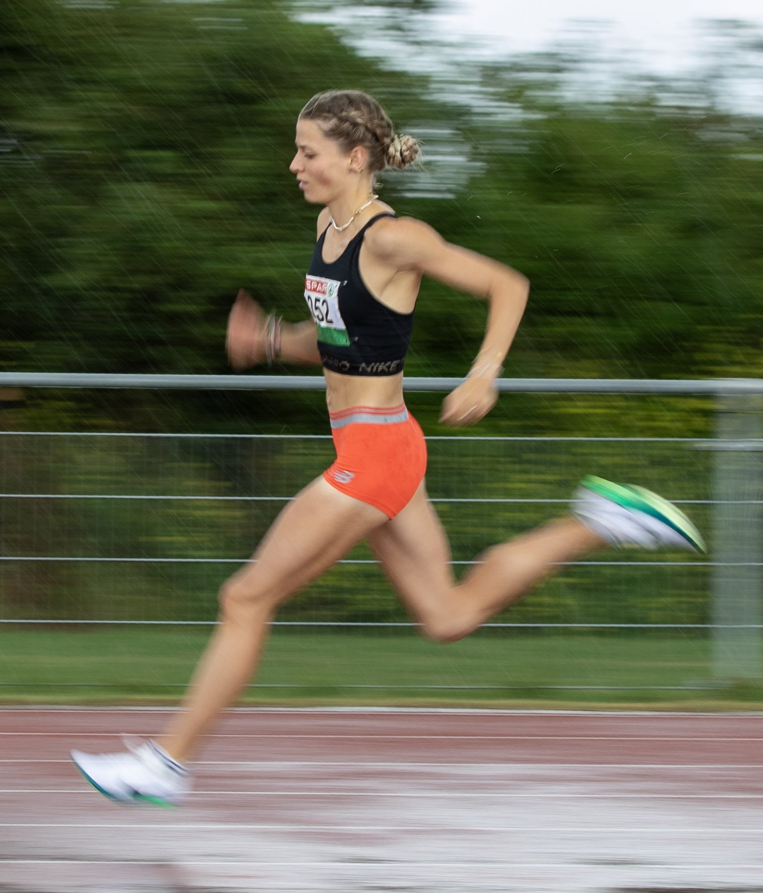 A female athlete running on a track during daytime, wearing a black sports top, orange shorts, and white running shoes, with a race number bib attached to her top.