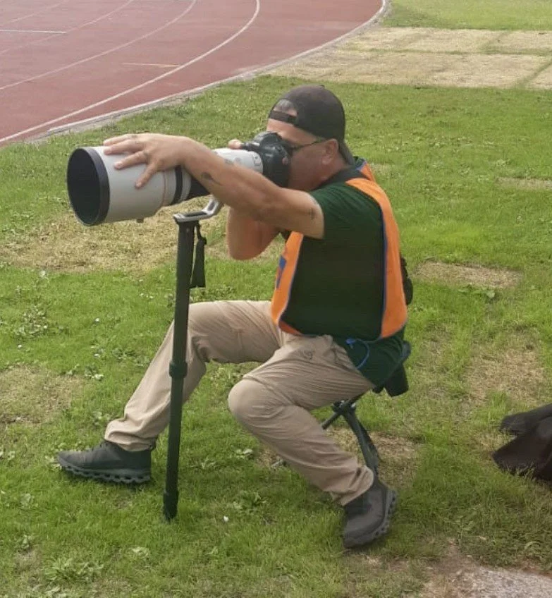 A man is sitting outdoors on a small foldable chair on grass, aiming a large telescope in a seated position near a running track.