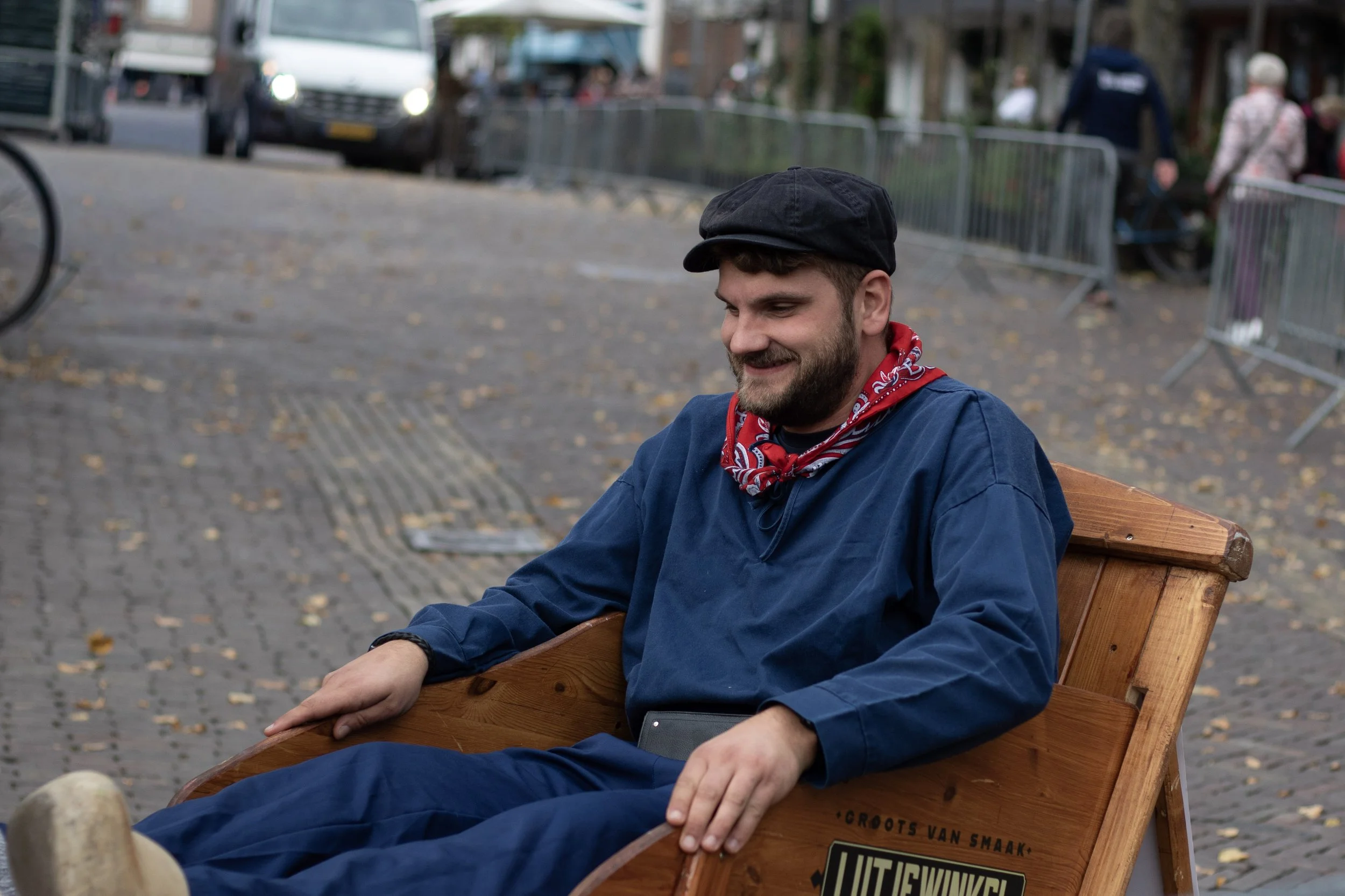 A man with a beard and mustache wearing a black cap, red bandana, and blue outfit sitting on a wooden bench in an outdoor setting that looks like a street or park with people, cars, and barricades in the background.