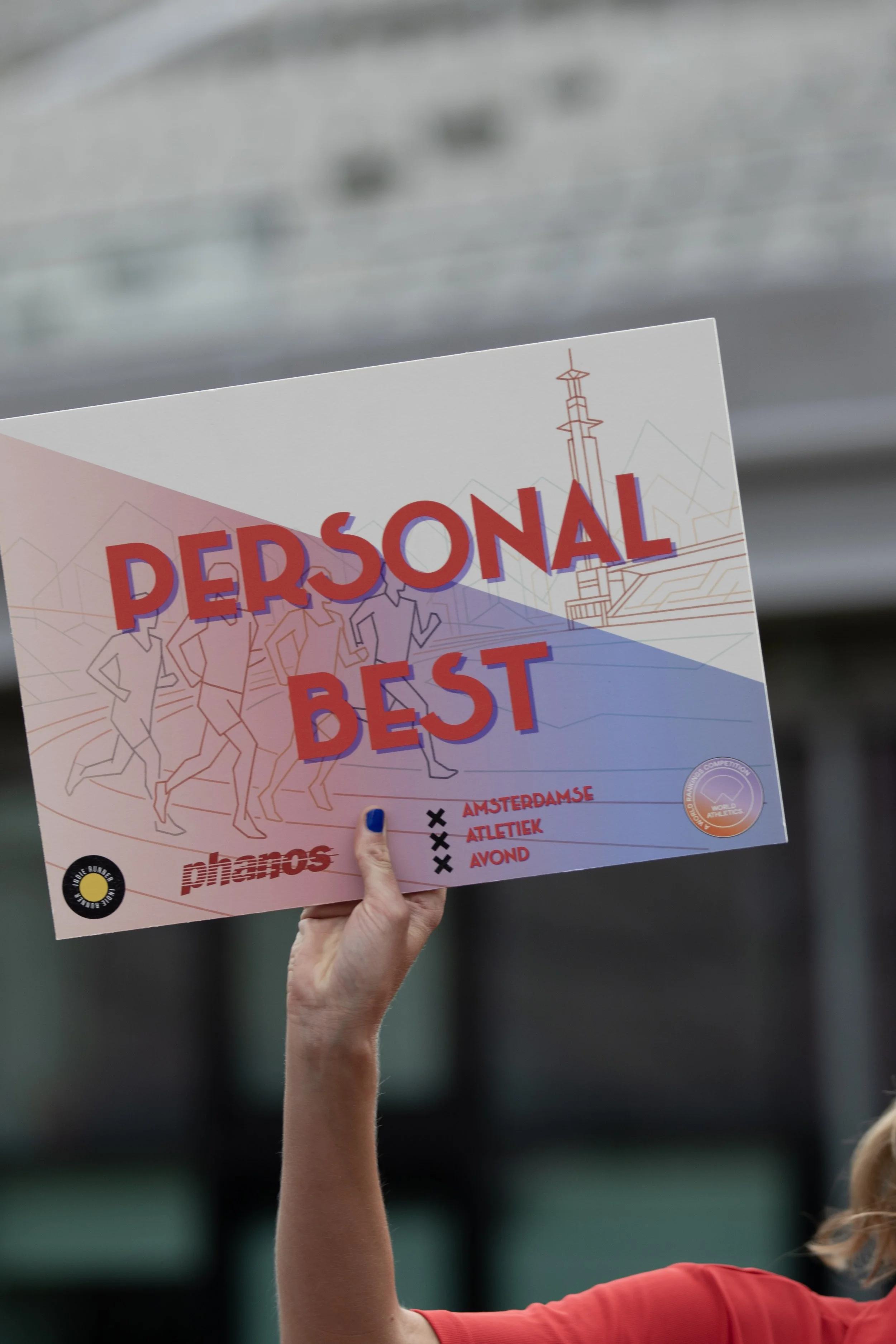 A person with blue nail polish is holding a sign that reads "Personal Best" during an athletic event, with illustrations of runners and a tower in the background.