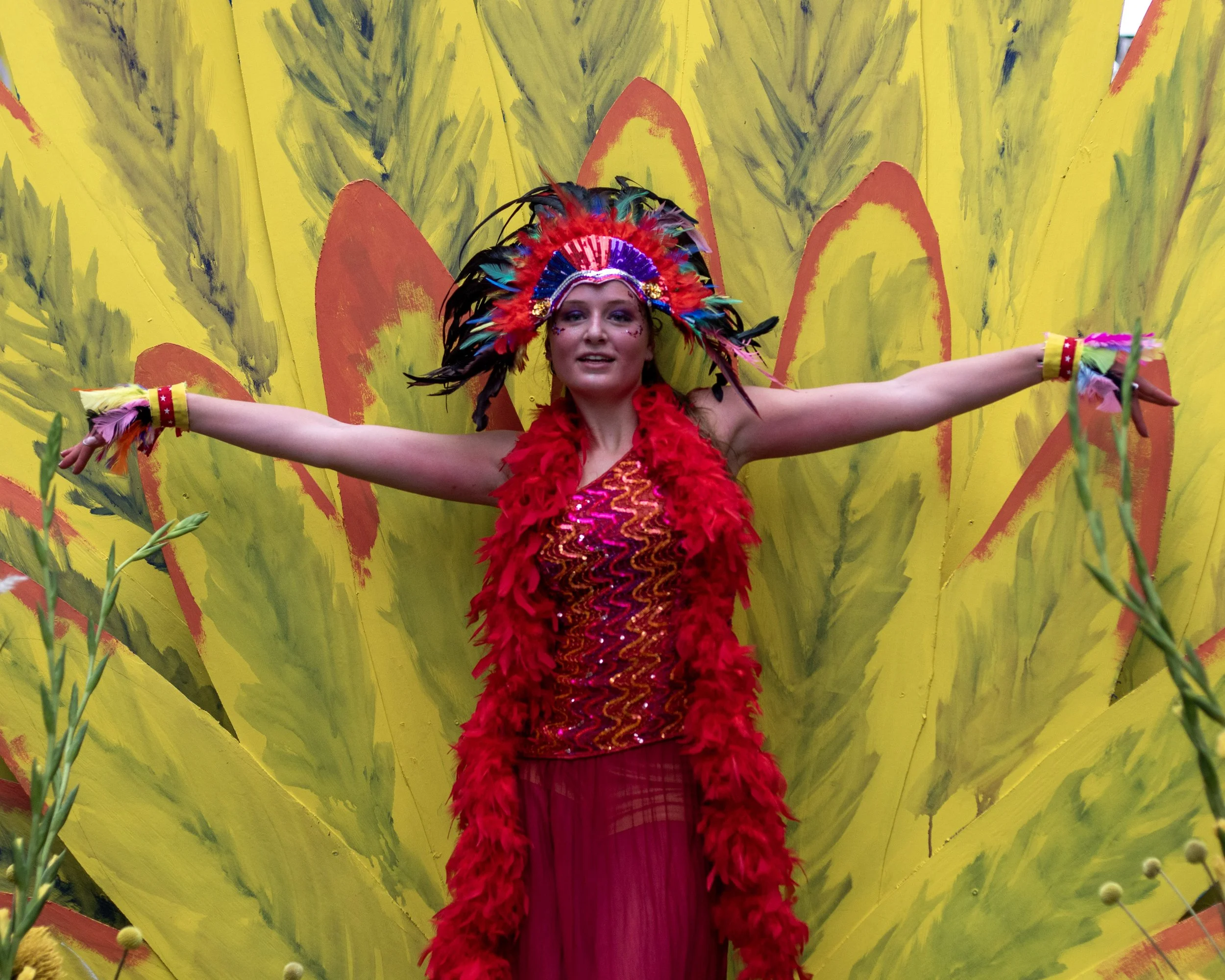A woman dressed in a colorful, feathered headdress and red costume stands with arms outstretched in front of a painted yellow backdrop with red and green accents, seemingly performing or celebrating.