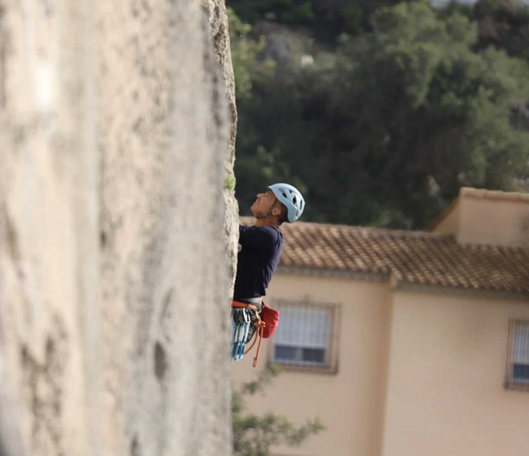 A person rock climbing outdoors, wearing a helmet and climbing gear, with a house and trees in the background.