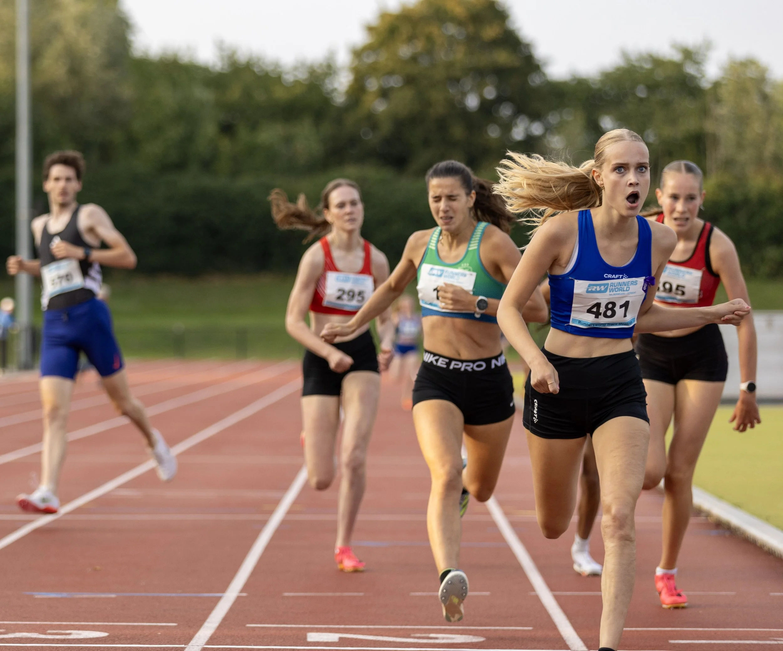 A group of female sprinters competing in a race on a red track field, with some women visibly exerting effort as they run, while one runner in front appears to be leading.