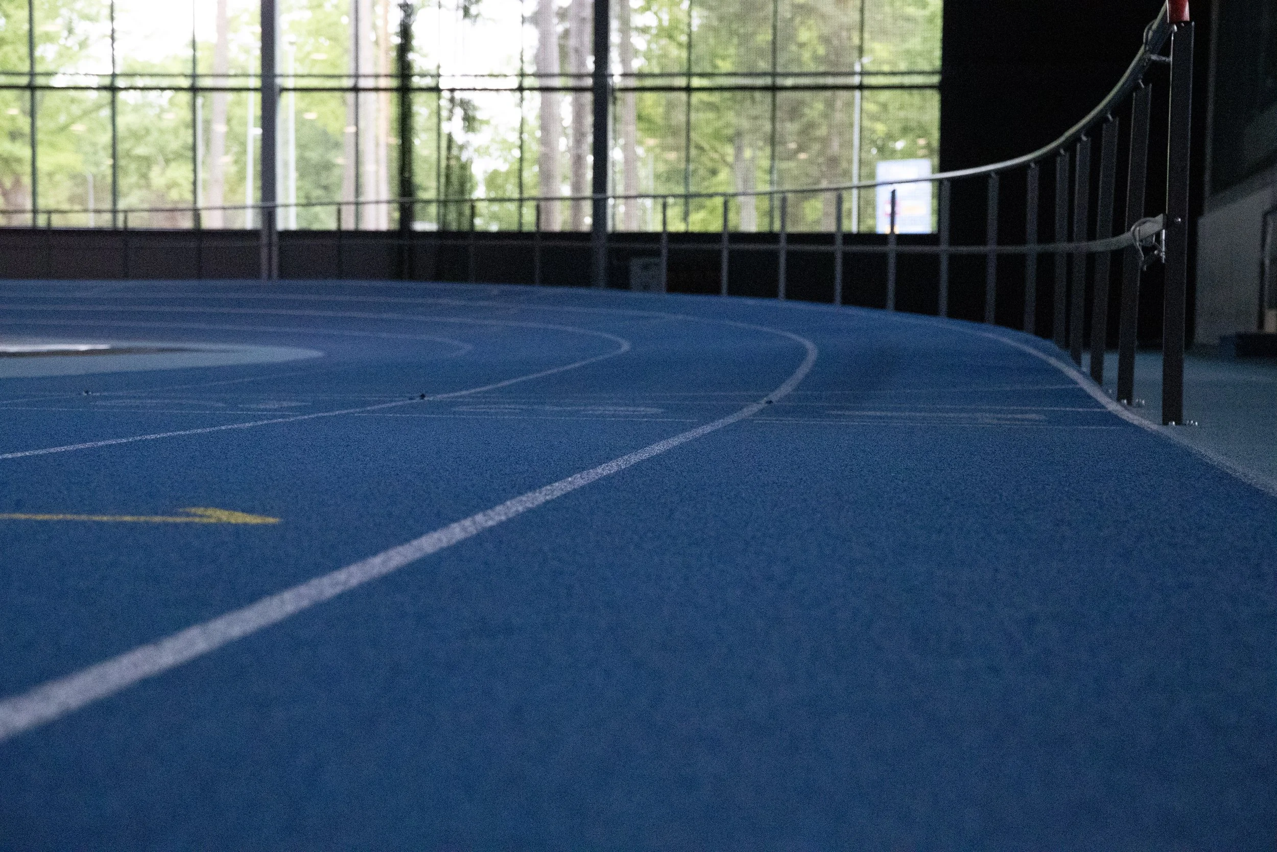Indoor athletic track with blue surface, surrounded by large windows showing green trees outside.