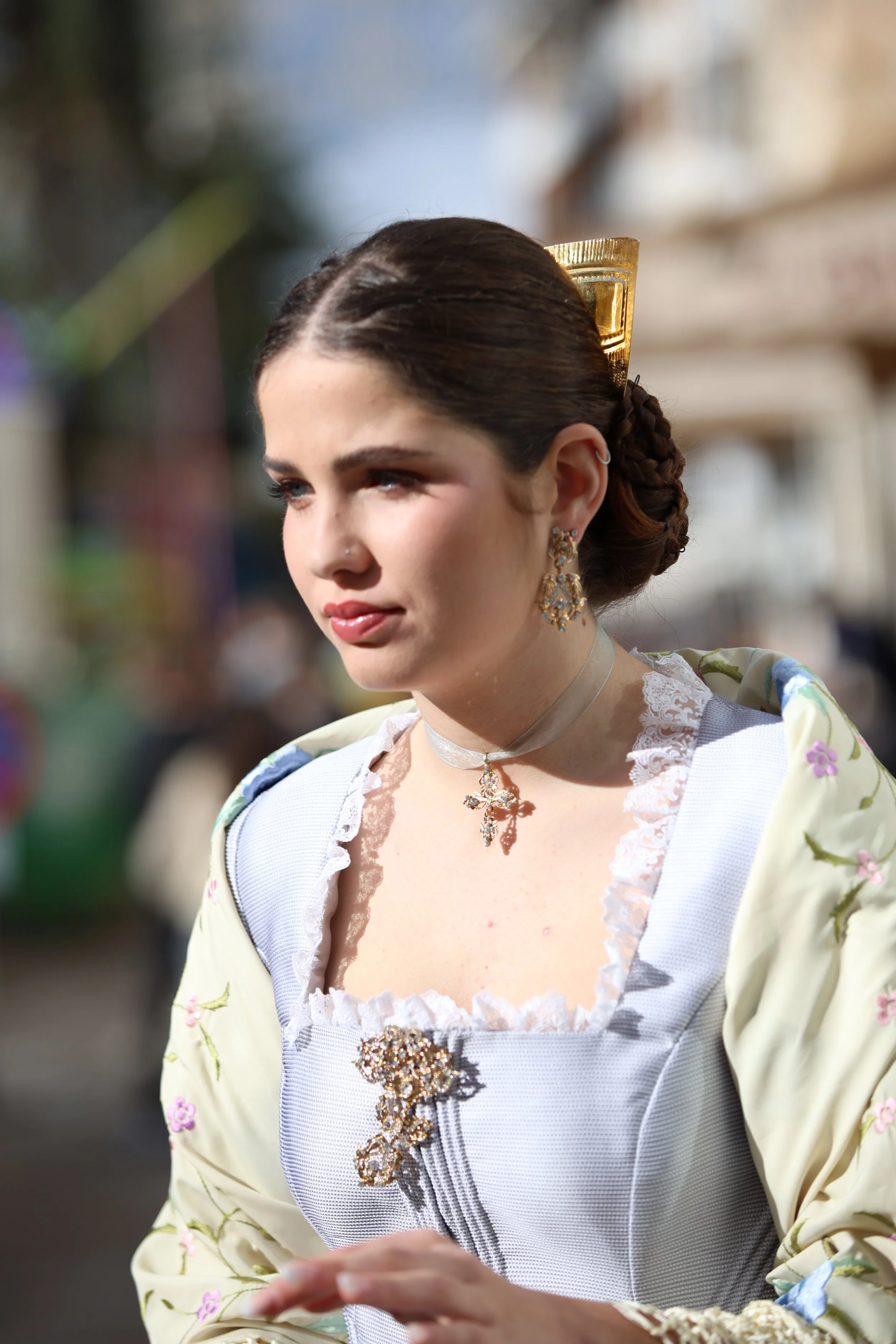 A young woman wearing large tortoiseshell sunglasses and earrings outdoors on a sunny day, with a blurred background of trees and buildings.