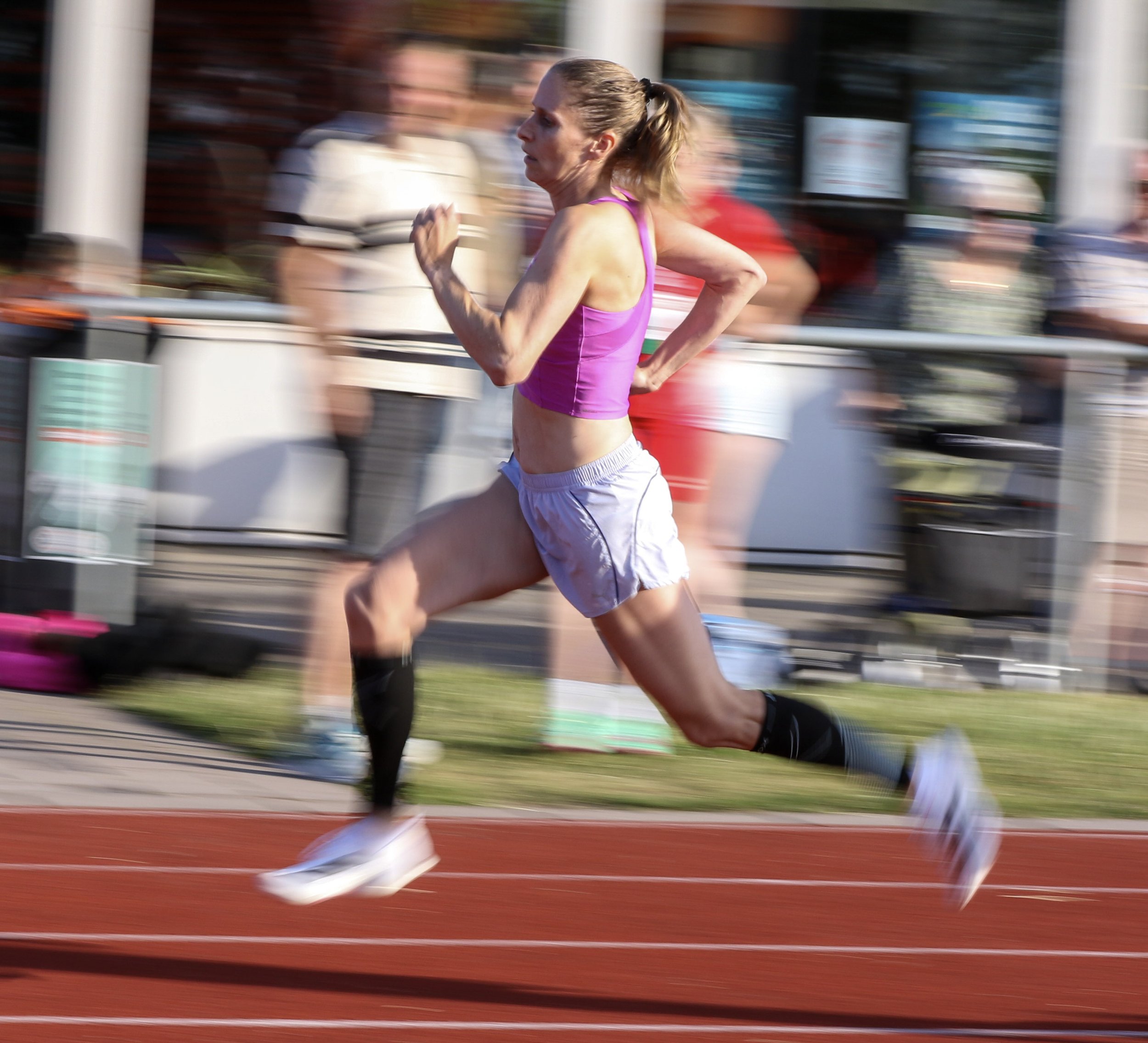 A woman running on a track during a race, wearing a purple sports bra, white shorts, black knee-high socks, and white running shoes.