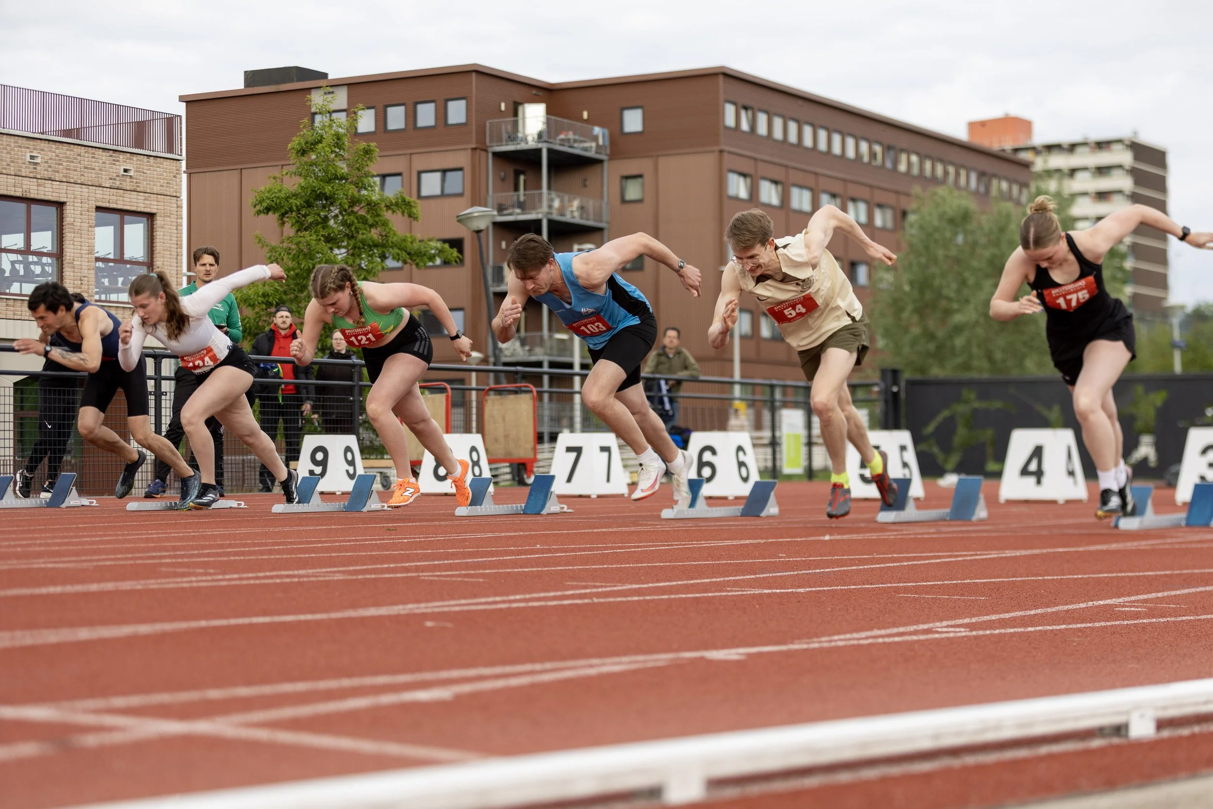 Athletes at the starting line of a race on a red track, with a brown building and trees in the background.