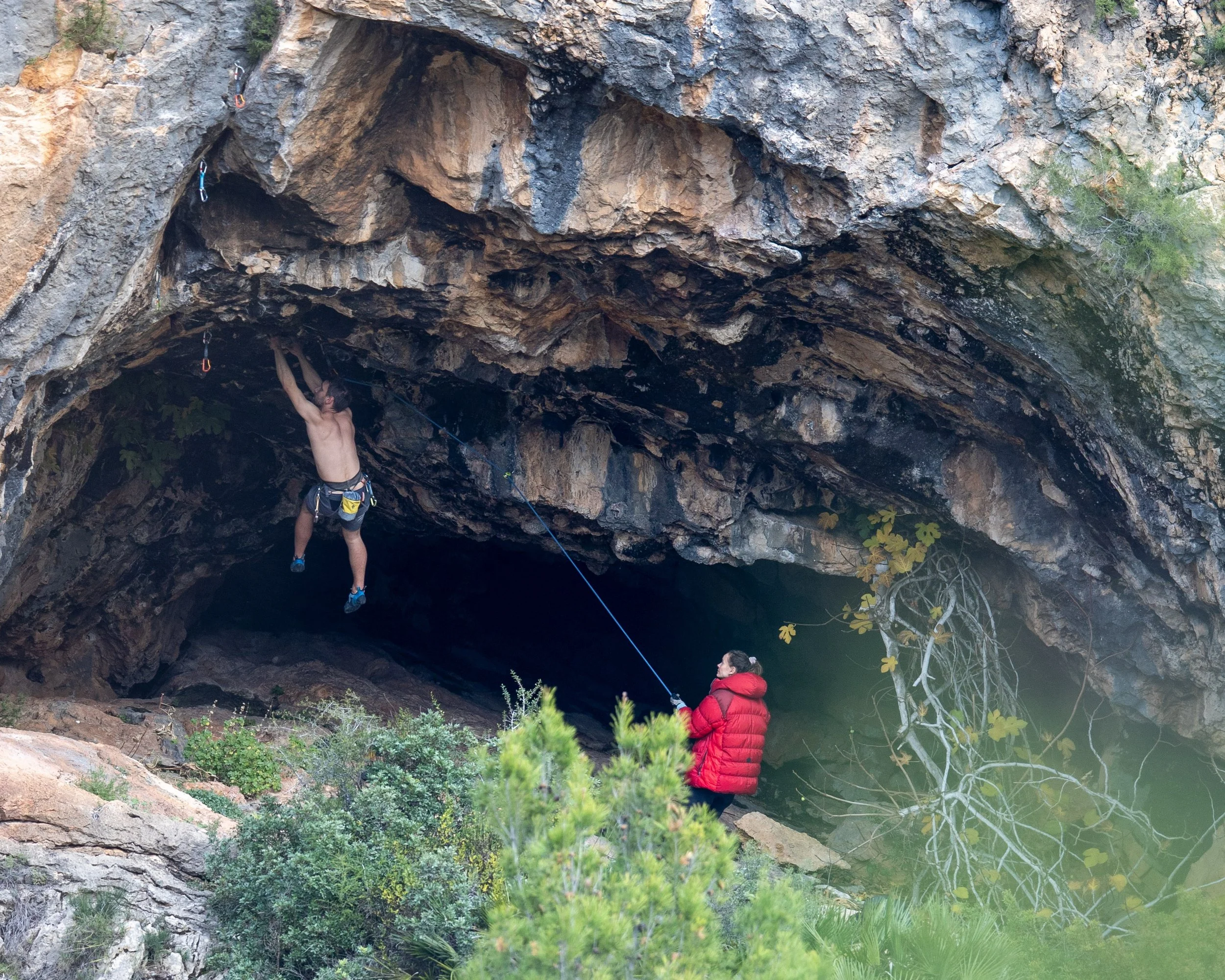 Two people rock climbing on a rock formation near a body of water, with one person climbing and another person monitoring from below, surrounded by natural vegetation.