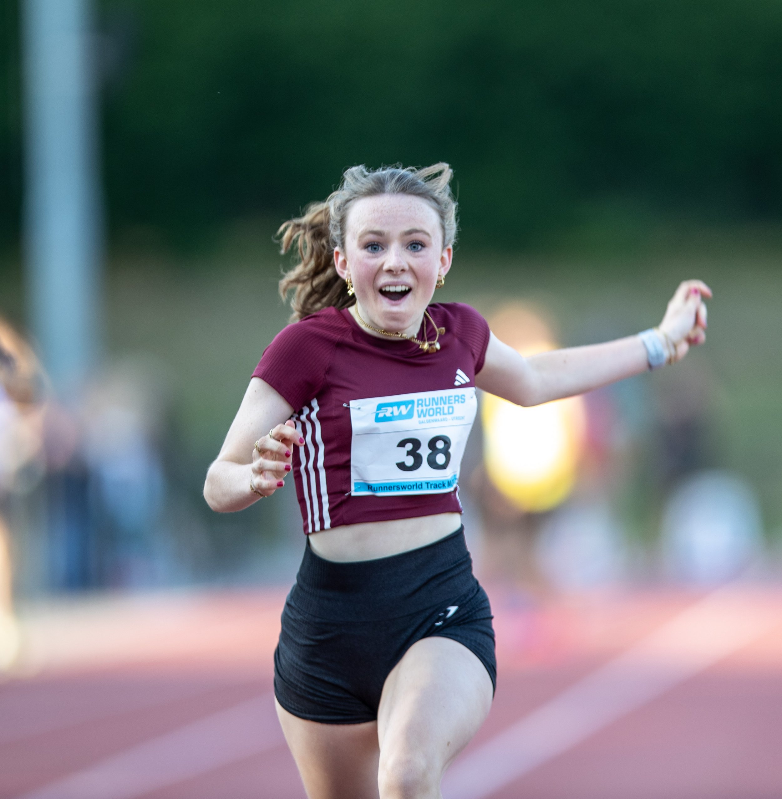 Young female athlete running on a track during a race, wearing a maroon athletic shirt with the number 38, black shorts, and gold jewelry, with a surprised or happy expression.
