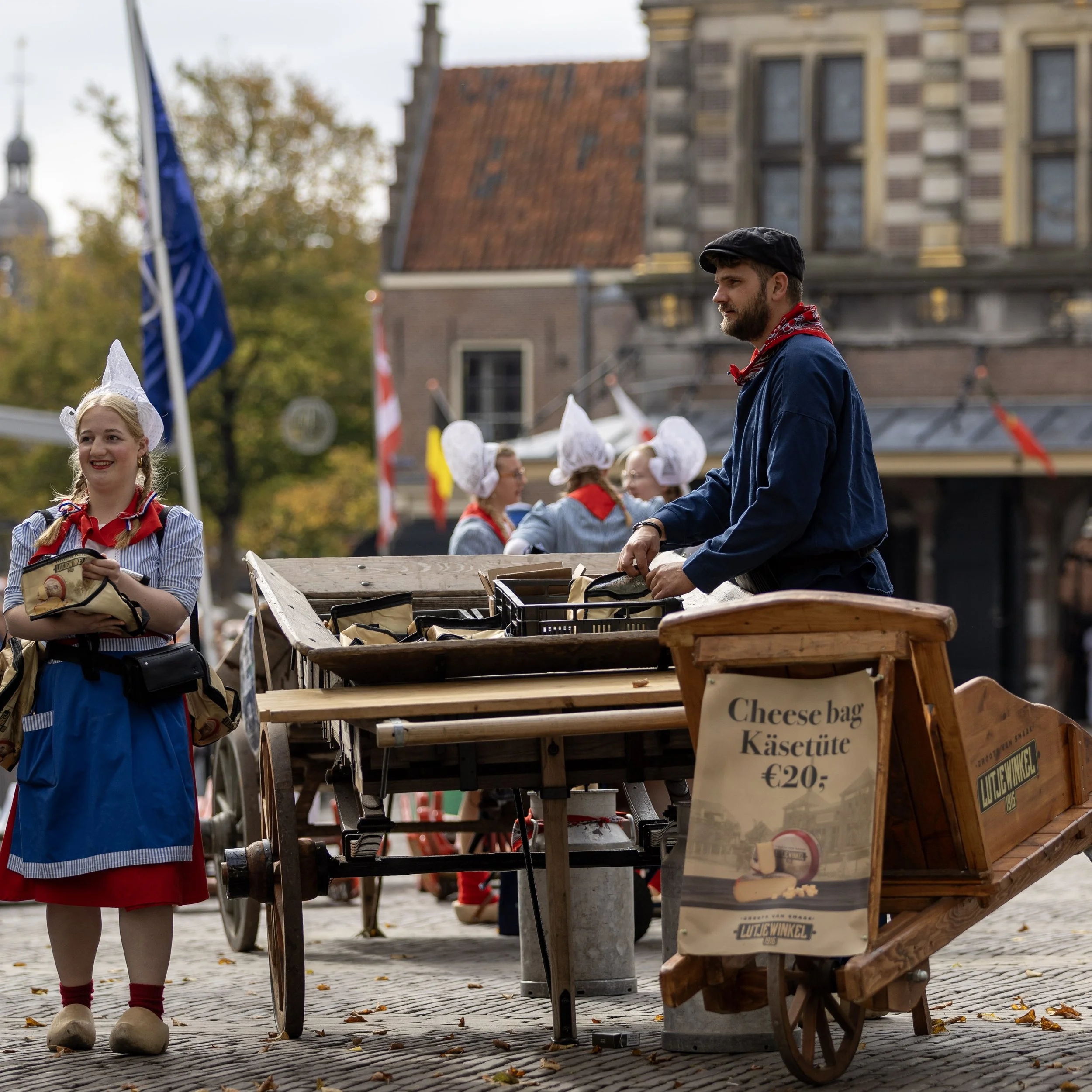 A man selling cheese bags at a market stall with a sign indicating 20 euros. Several women dressed in traditional European folk costumes with white head coverings and red neckerchiefs are in the background. The setting appears to be in a European town square during autumn.