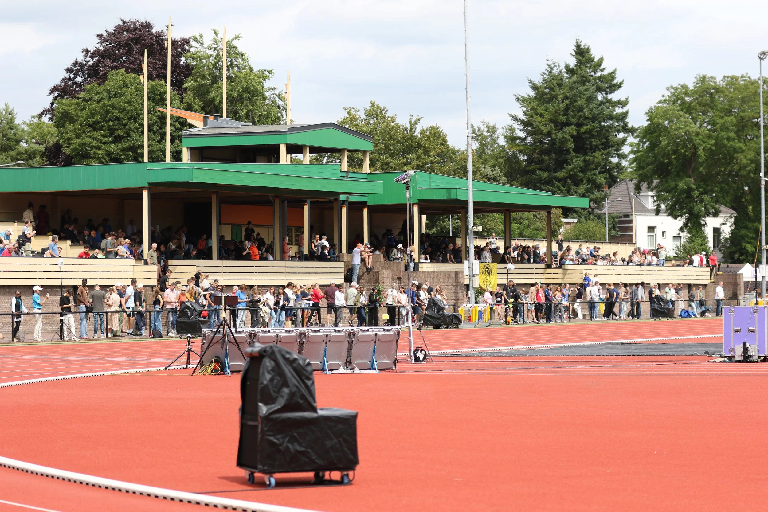 Crowd of people waiting in line at a sports stadium with a red running track in the foreground and a green grandstand with trees in the background.