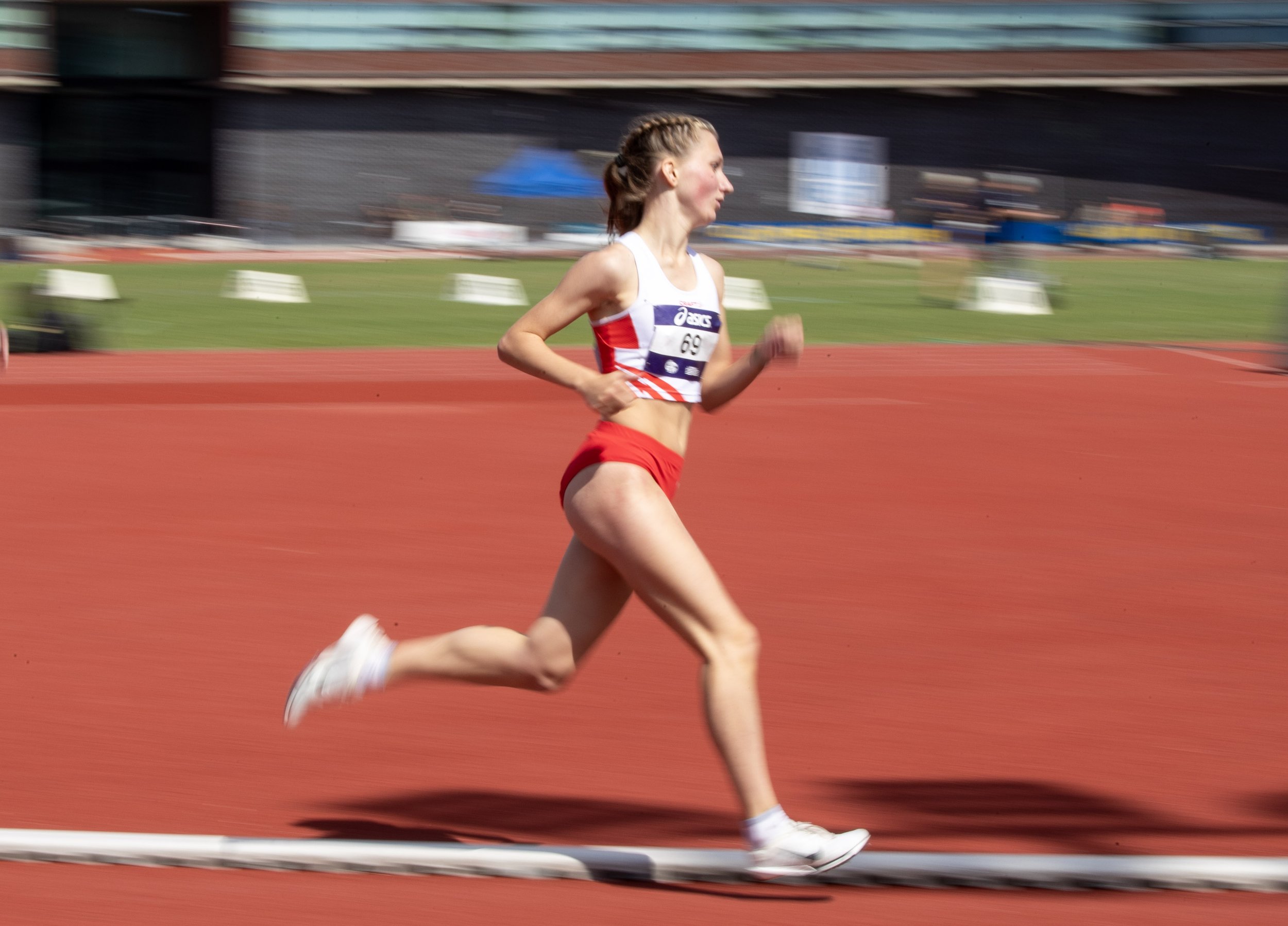 A female athlete running on a red outdoor track during a race, wearing a red and white athletic outfit with a bib number 69, with a blurred background of the stadium and spectators.