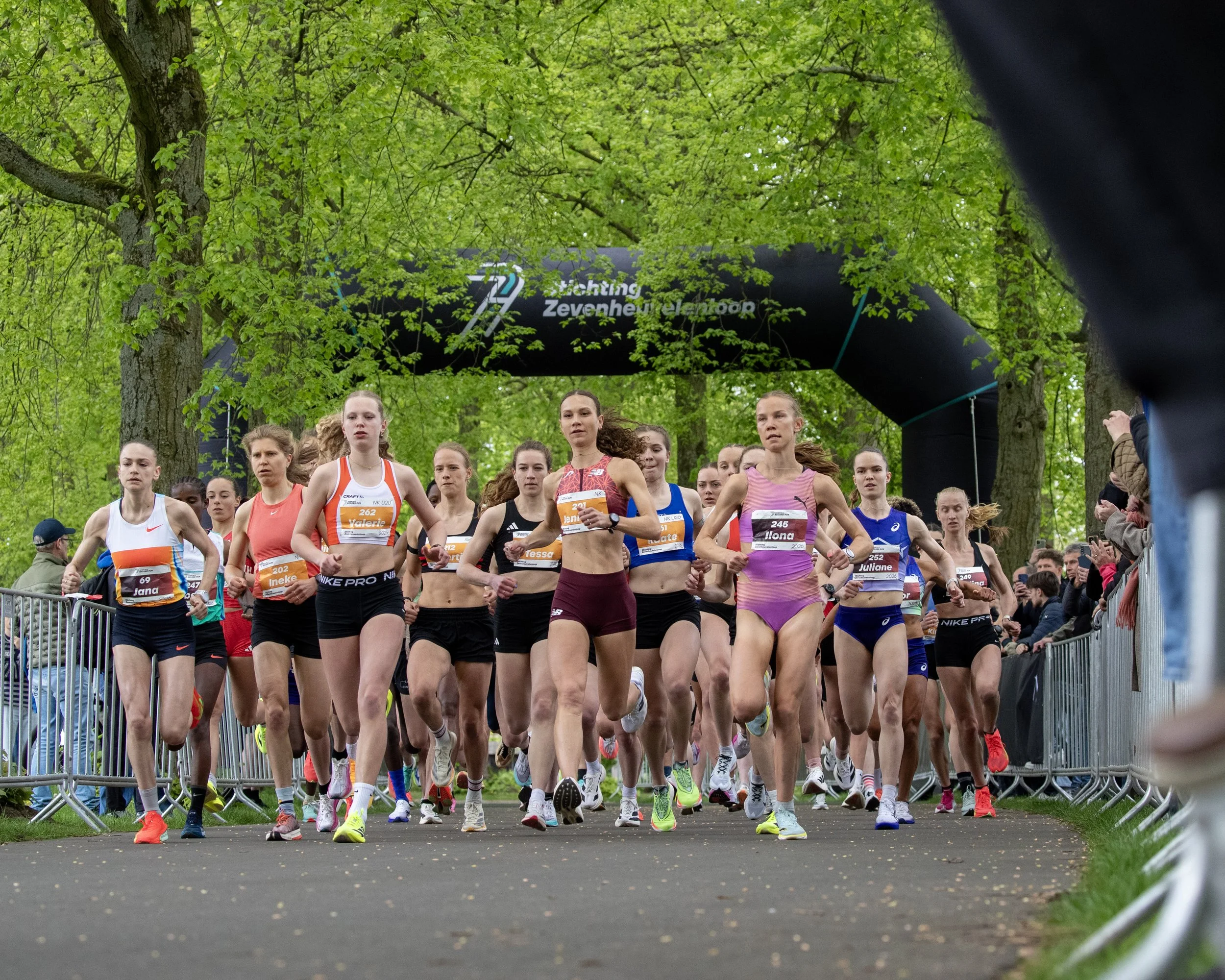 Group of female runners at the start of a race in a park with green trees and a black inflatable archway overhead.