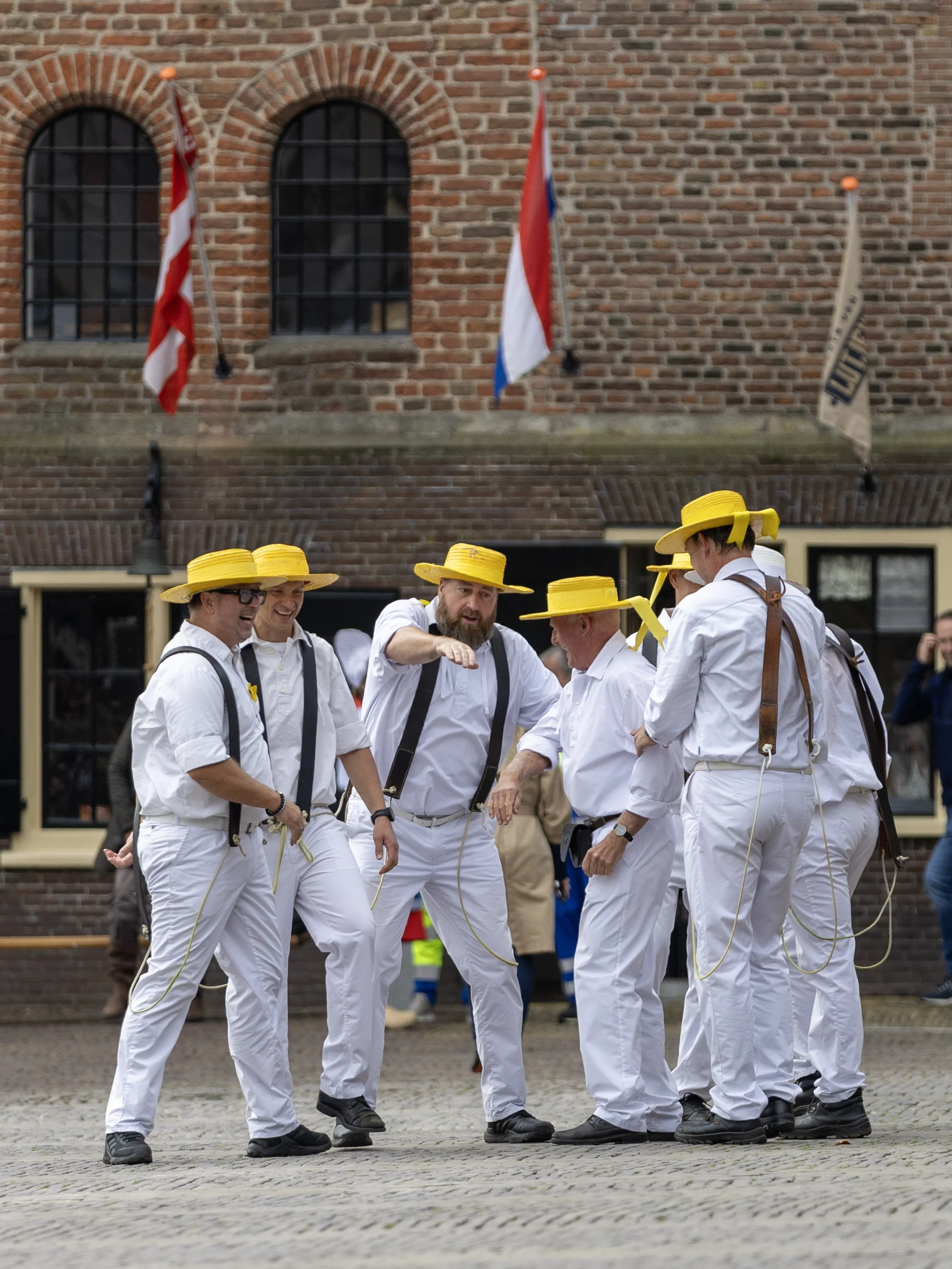 Group of men dressed in white shirts and pants, wearing yellow hats, standing in a circle on a cobblestone street, engaged in conversation or a performance. Background features a brick building with three windows and flags.