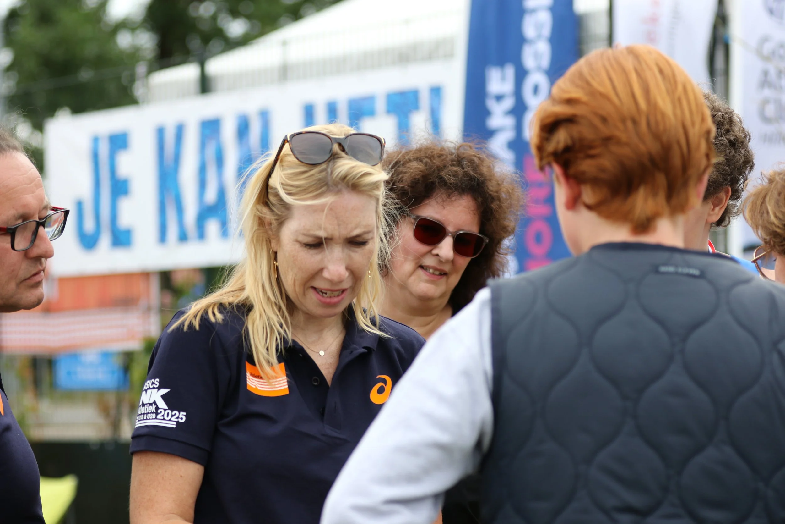 Group of people gathered outside, with a woman wearing a navy polo shirt with logos, sunglasses on her head, and blonde hair, talking to a person with red hair from behind, and others around them, during an outdoor event or gathering.