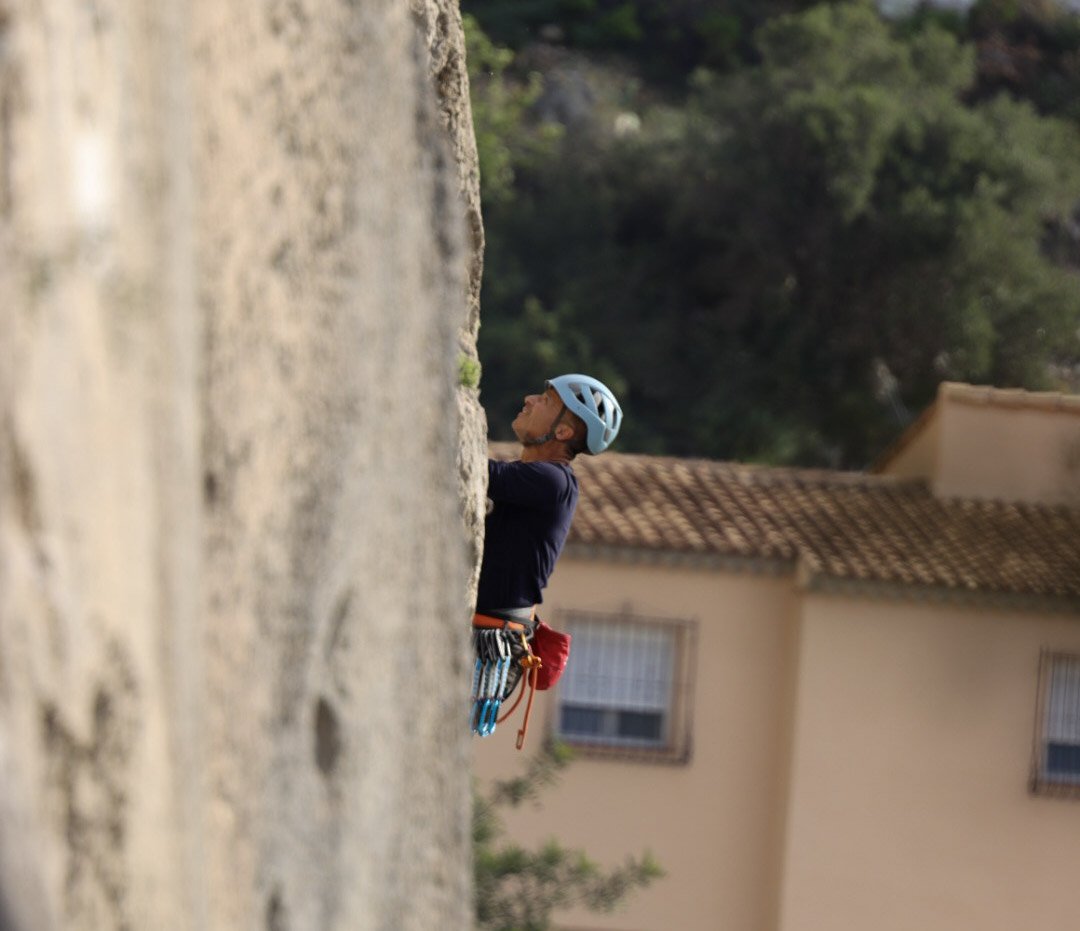A person wearing a blue helmet and dark clothing climbing a rock face, with climbing gear attached to their harness, against a background of trees and a building with a tiled roof.