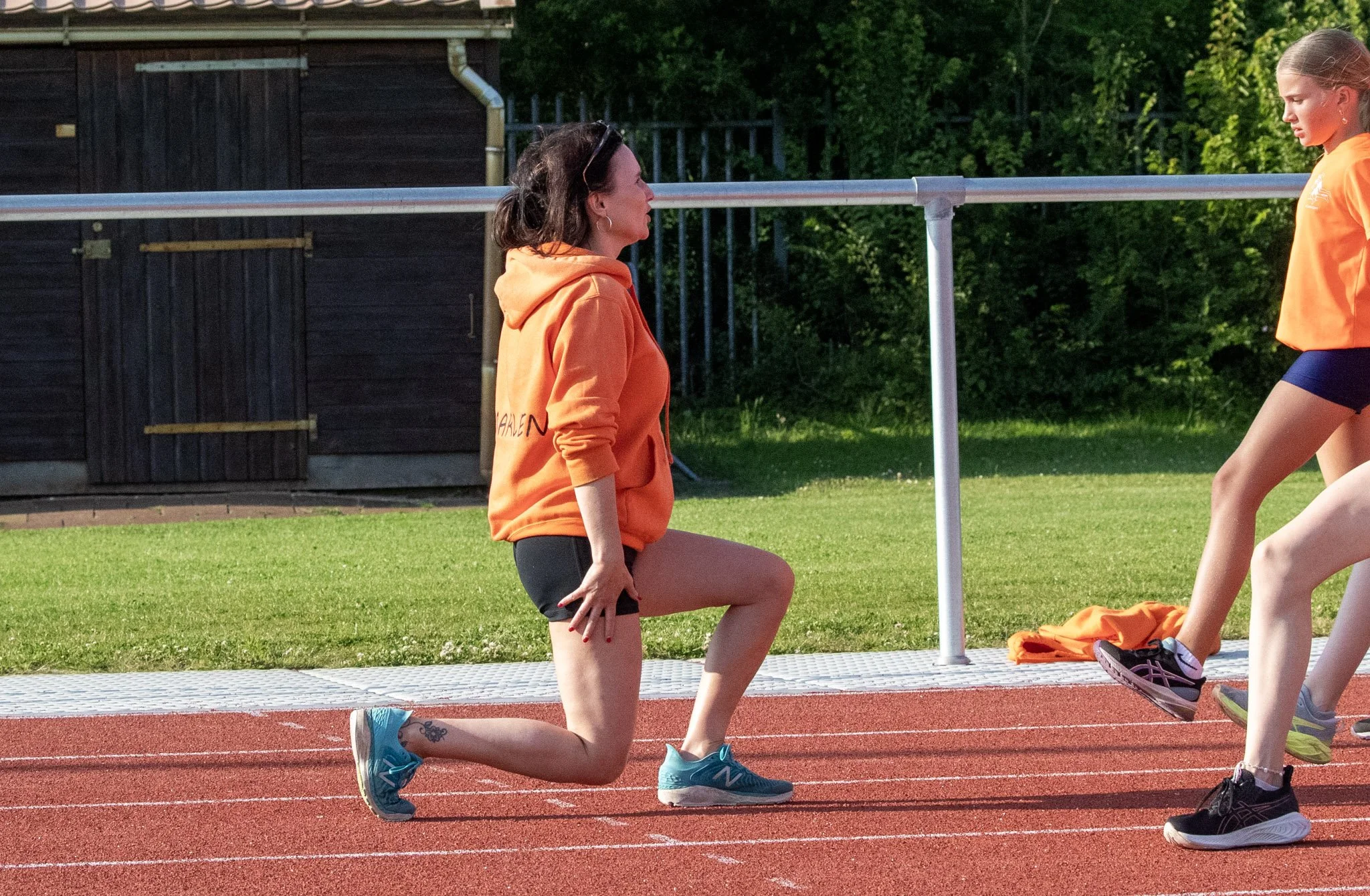 A woman in a bright orange hoodie and black shorts is kneeling on a running track with one knee on the ground and one foot on the track. She appears to be preparing for or participating in a workout or race. There is a girl in similar orange athletic attire standing a few feet away, on a sunny day with green trees in the background.