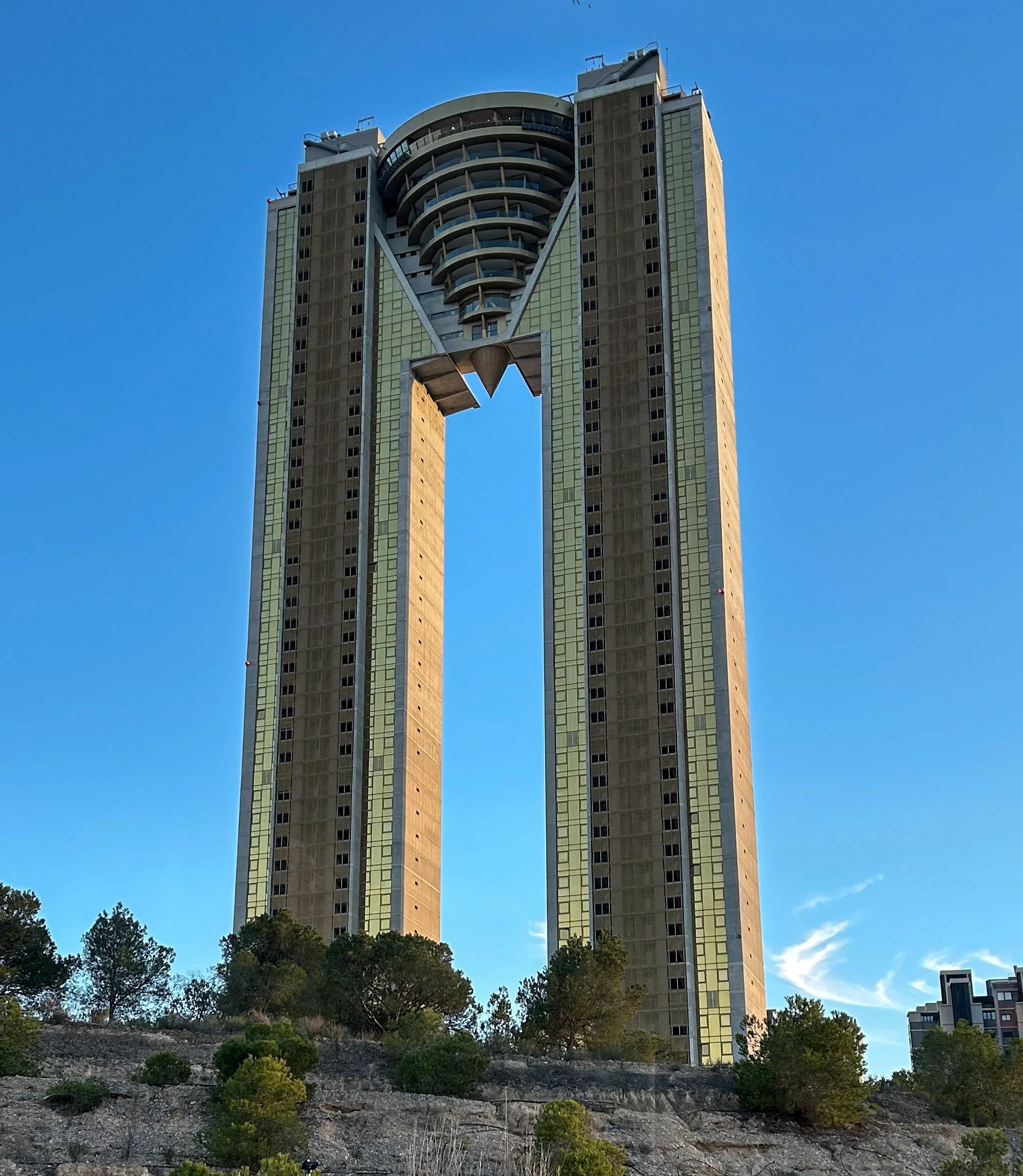 A tall modern skyscraper with a distinctive open square in the middle, situated on a hill with trees and bushes, under a clear blue sky.