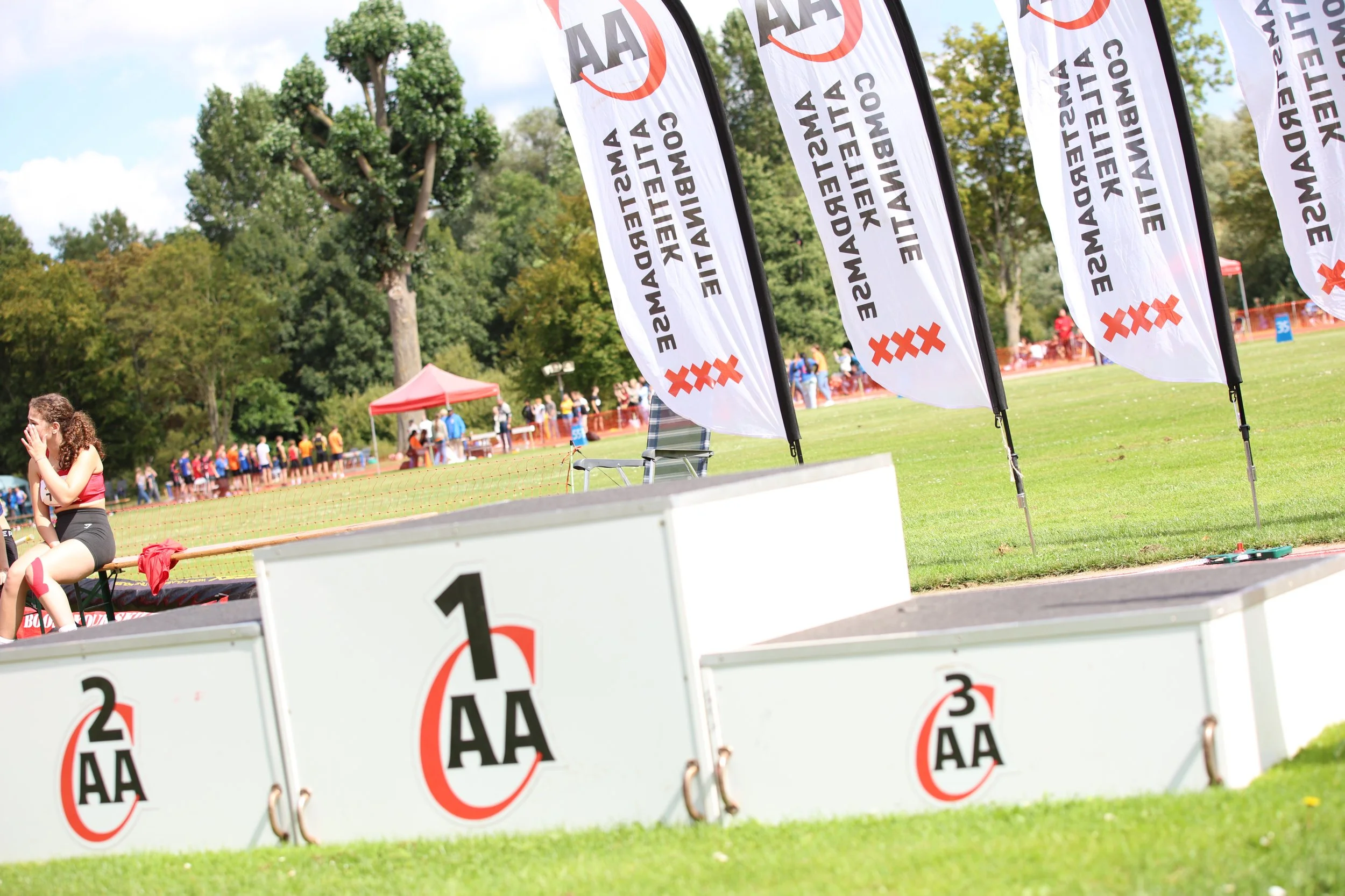 Athletic running track with flags bearing the AA Amateur Athletics Association logo, a woman sitting on the second-place podium, and a line of spectators in the background against a green park setting.