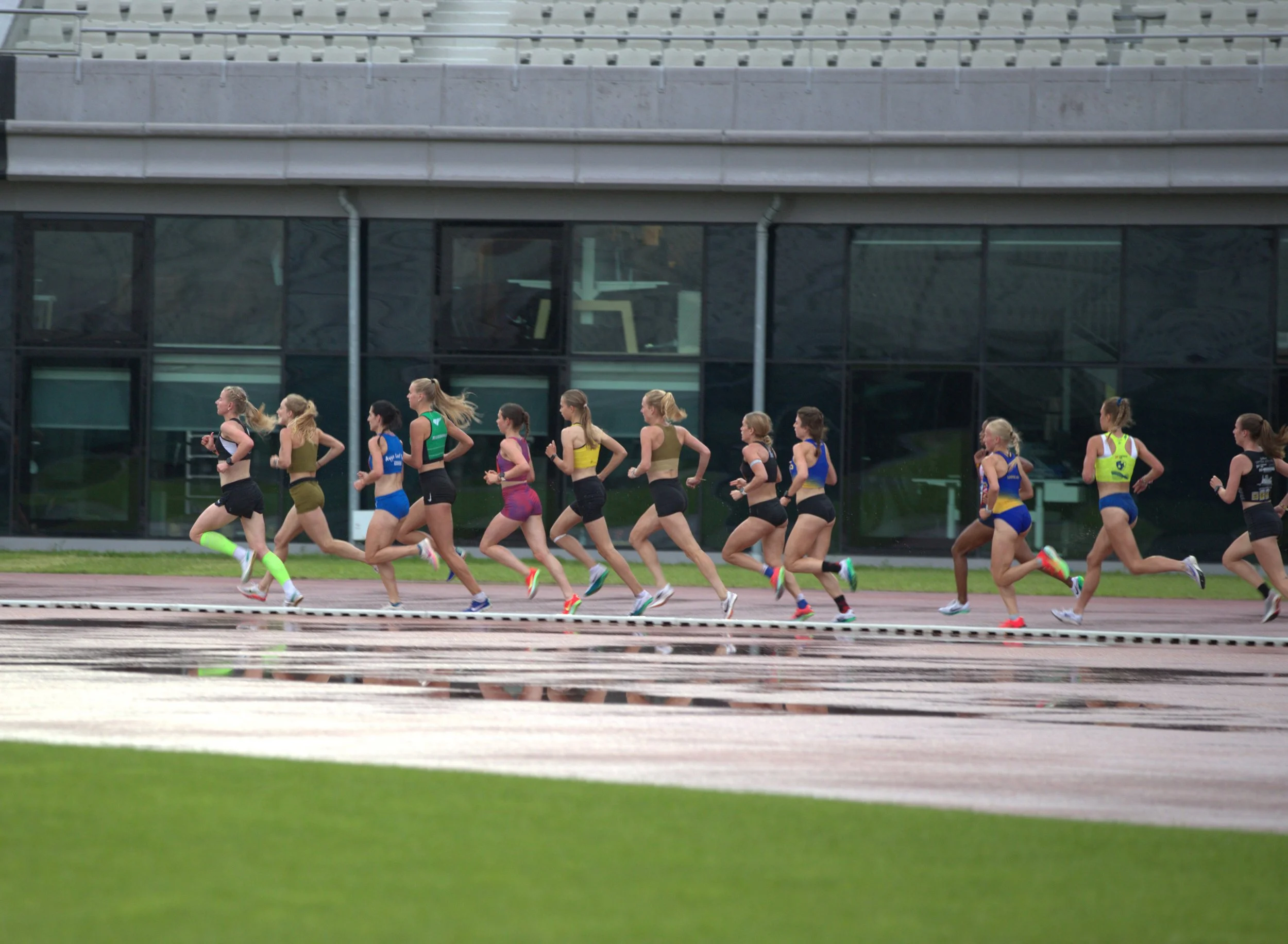 A group of female runners competing in a track race, running on a wet track with a modern building in the background.