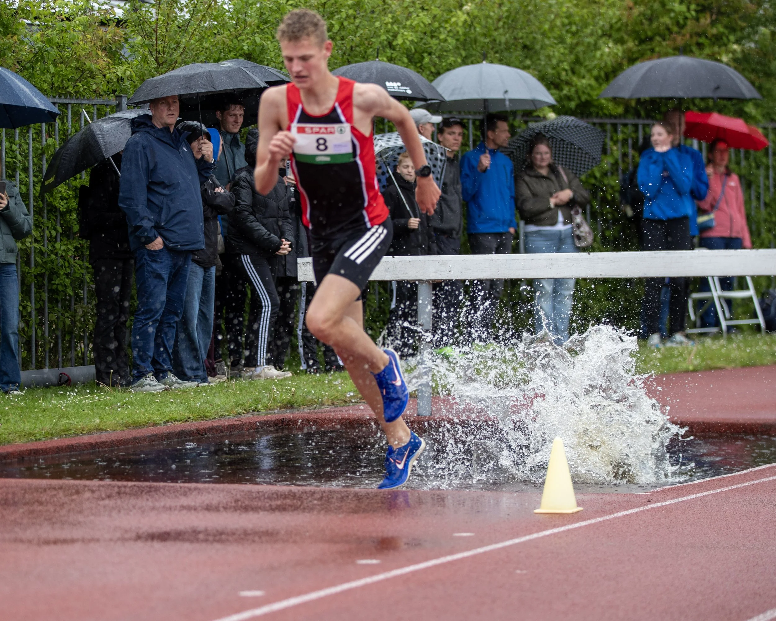 A male athlete participating in a steeplechase race jumps over a water obstacle on a red running track during a rainy day, with spectators holding umbrellas and watching along the fence.
