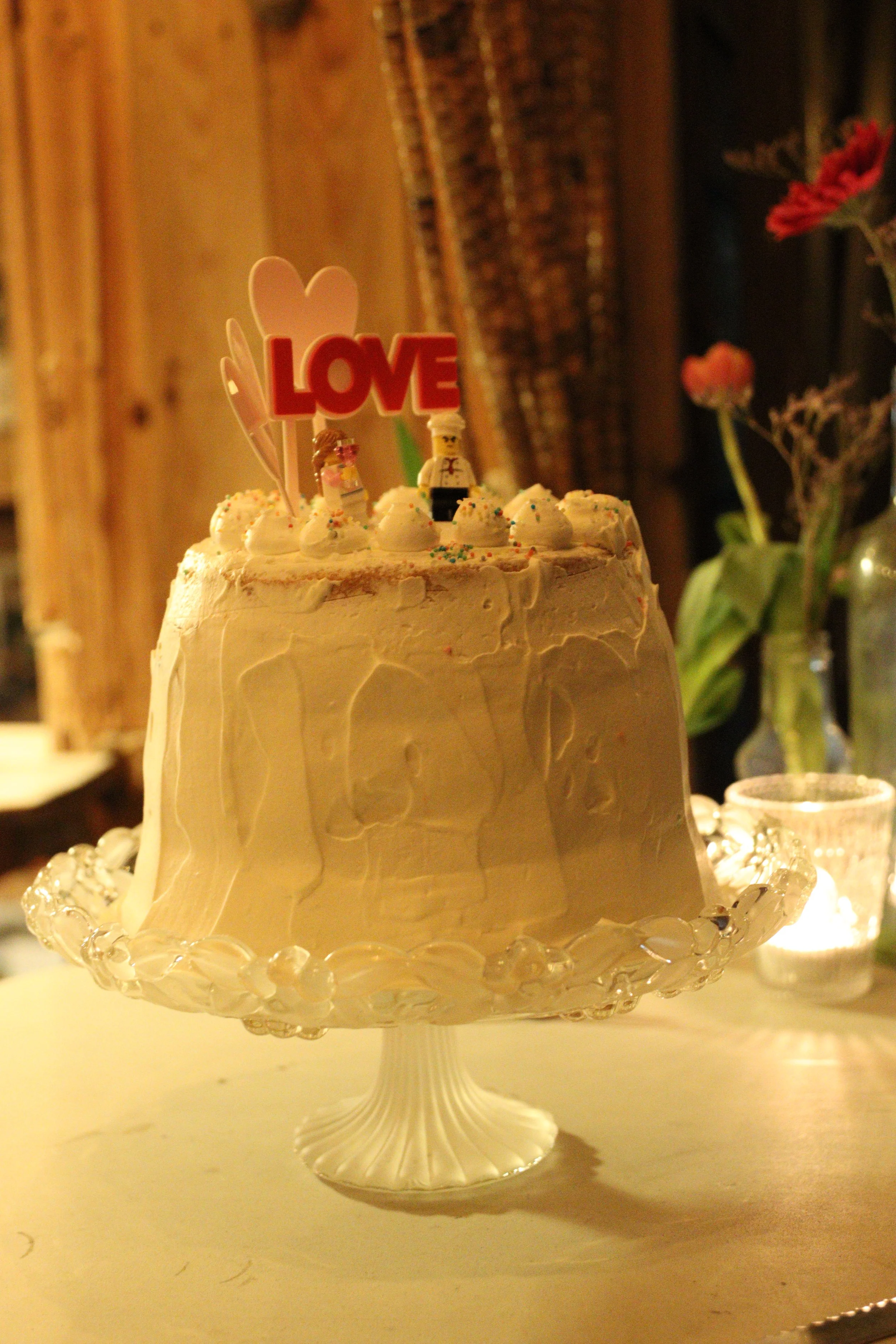 A decorated white wedding cake with small figures on top, including a 'LOVE' topper, placed on a glass cake stand with decorative edges, in a warmly lit setting with flowers and candles in the background.