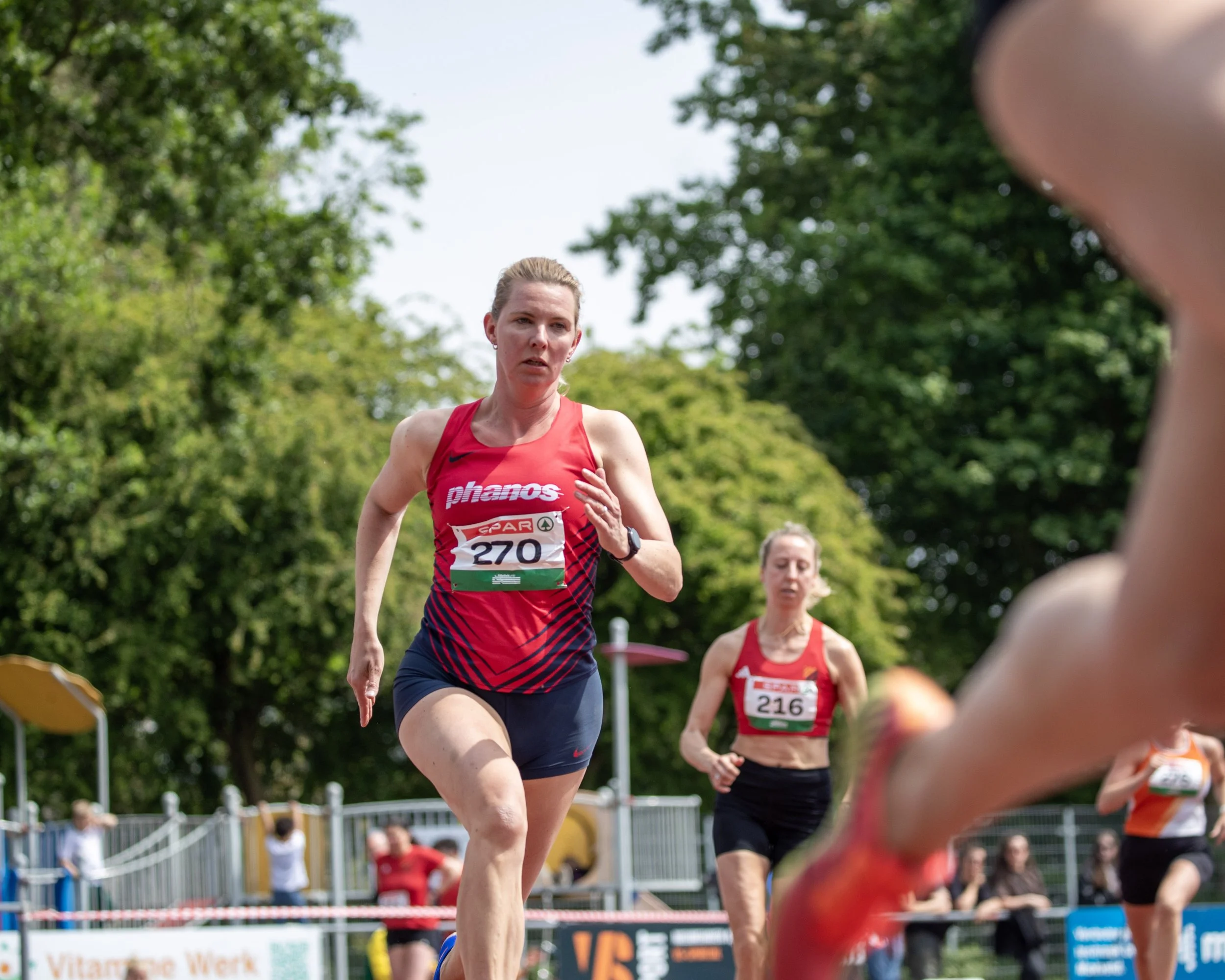 Female runner in a red tank top and black shorts participating in a race outdoors, with other runners and spectators in the background.