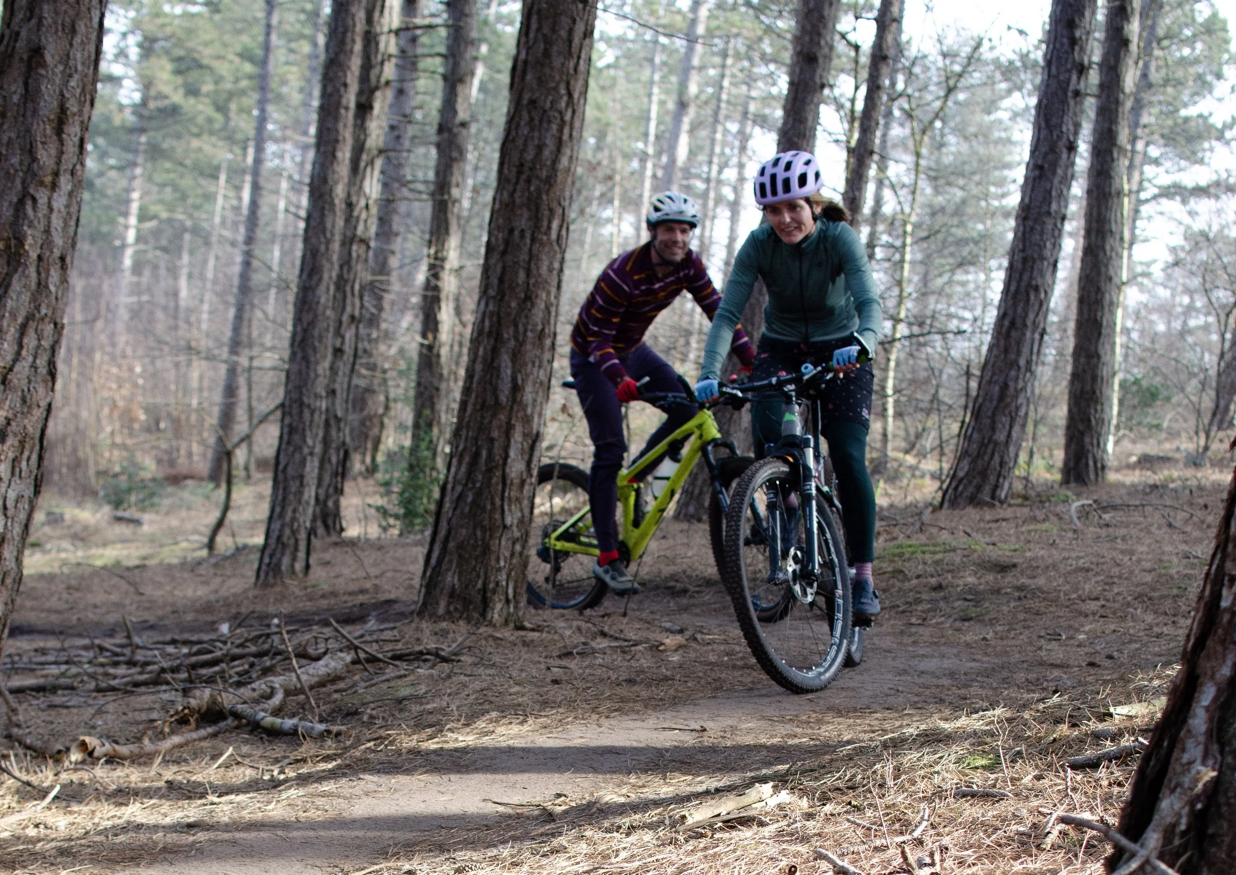 Two people mountain biking through a forest trail, wearing helmets and gloves, surrounded by tall trees.