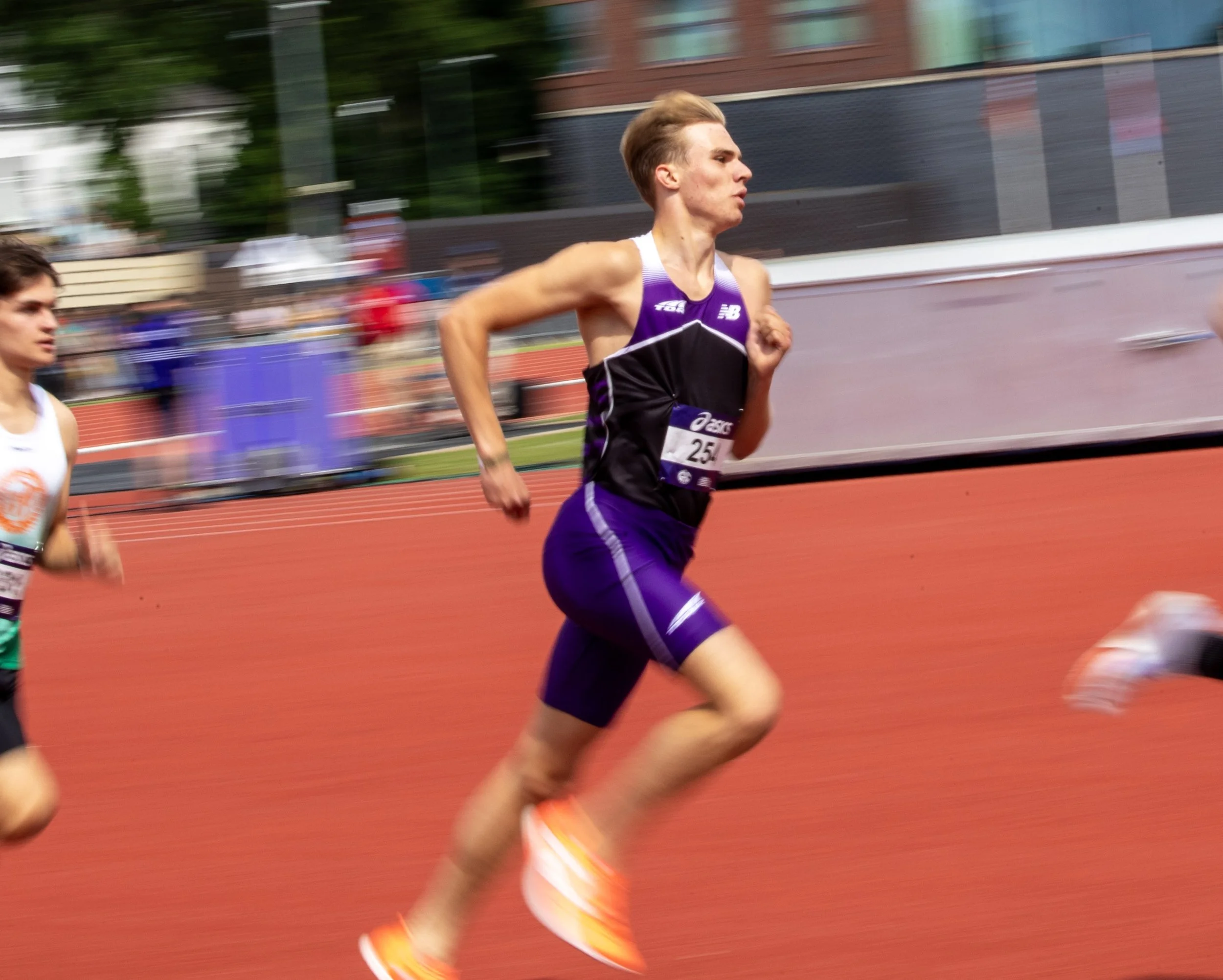 Young male athlete running on a red track during a race, wearing a purple and black athletic uniform and bright orange running shoes.