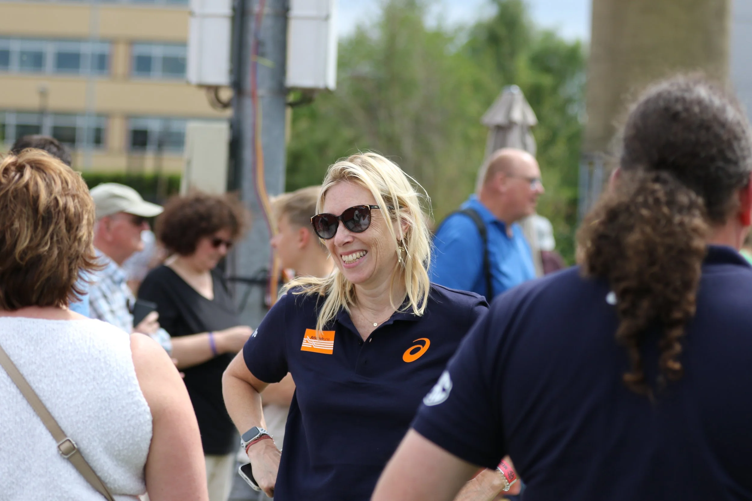 A group of people gathered outdoors, with a woman in the center wearing sunglasses and a navy shirt, smiling and engaging with others.