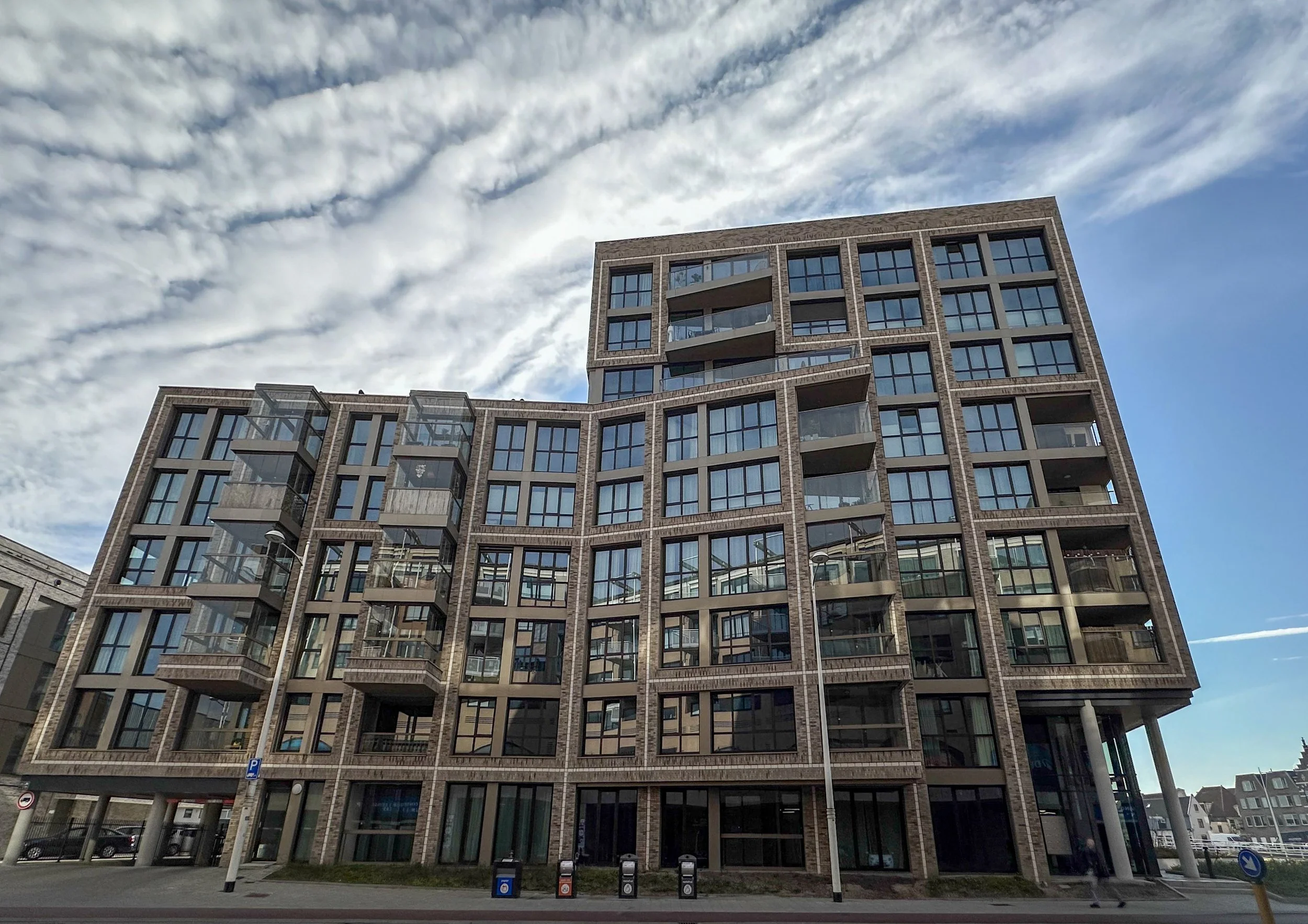 Modern multi-story residential building with a brick facade and large glass windows, situated on a city street under a partly cloudy sky.