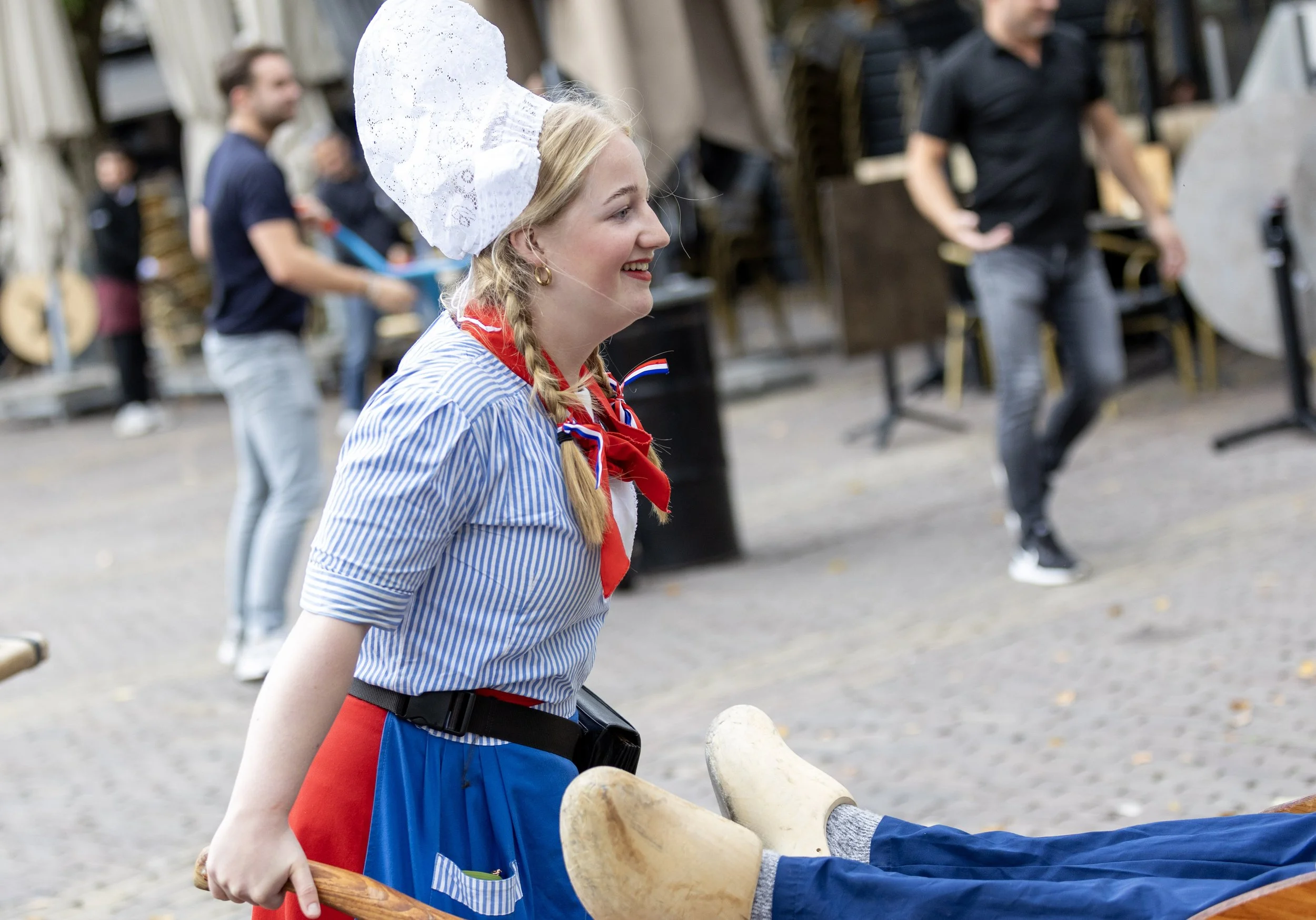 Young woman in traditional Dutch costume smiling, pushing a cart with a person’s legs in wooden shoes.