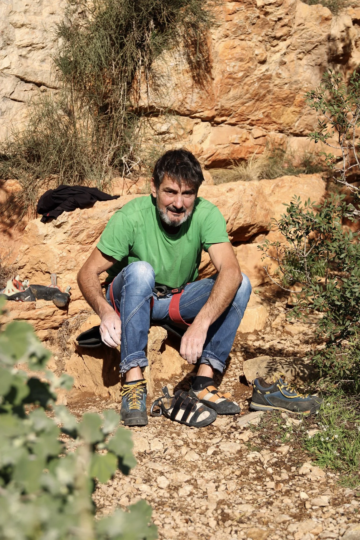 A man in a green shirt and jeans sits on rocky ground after rock climbing, with climbing shoes and outdoor gear around him, in a desert-like environment with orange rocks and sparse vegetation.