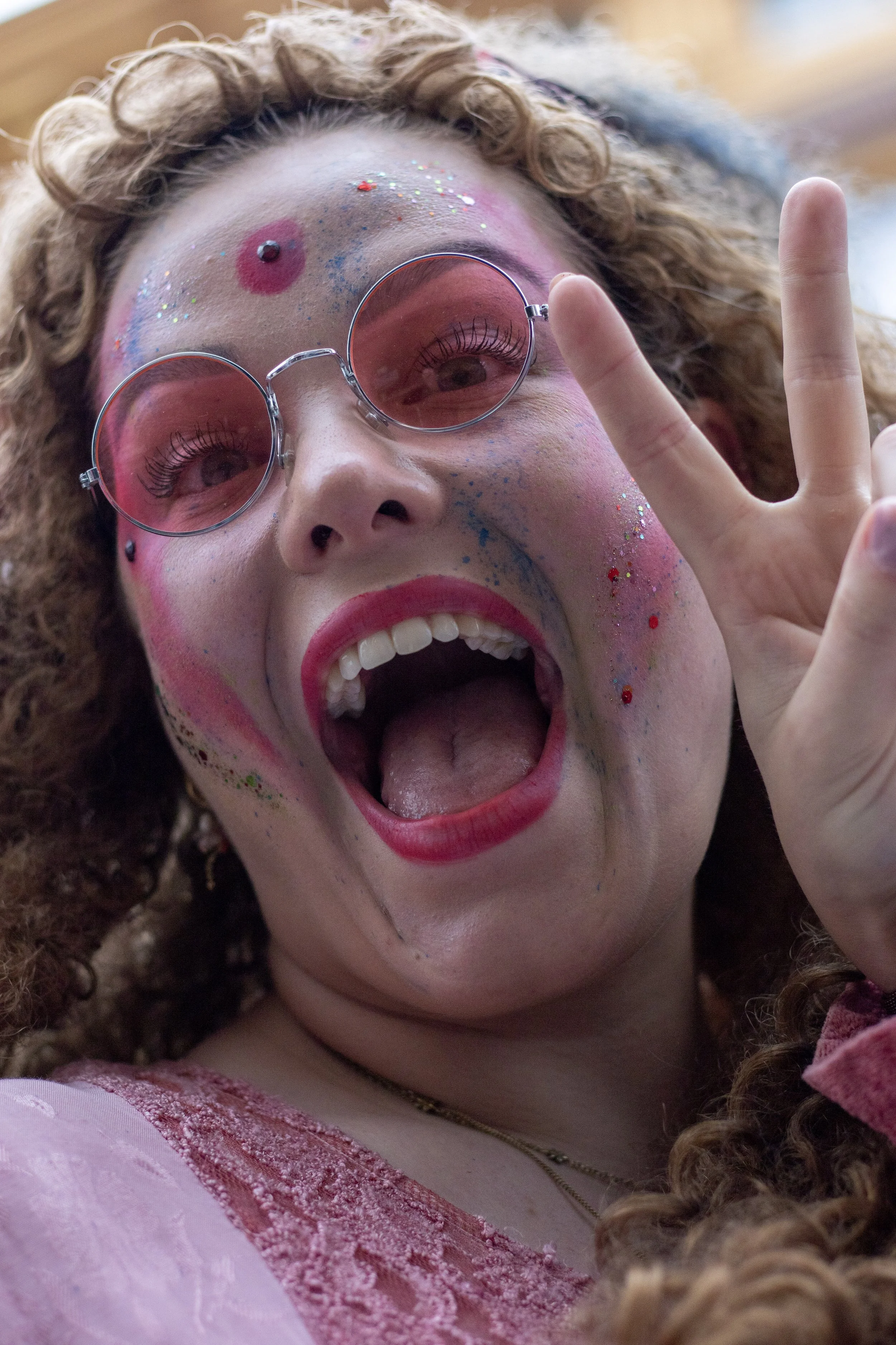 A young woman with curly hair and face paint in pink, blue, and glitter, wearing pink sunglasses, a pink dress with lace, and making a peace sign with her hand, smiling with her mouth open.