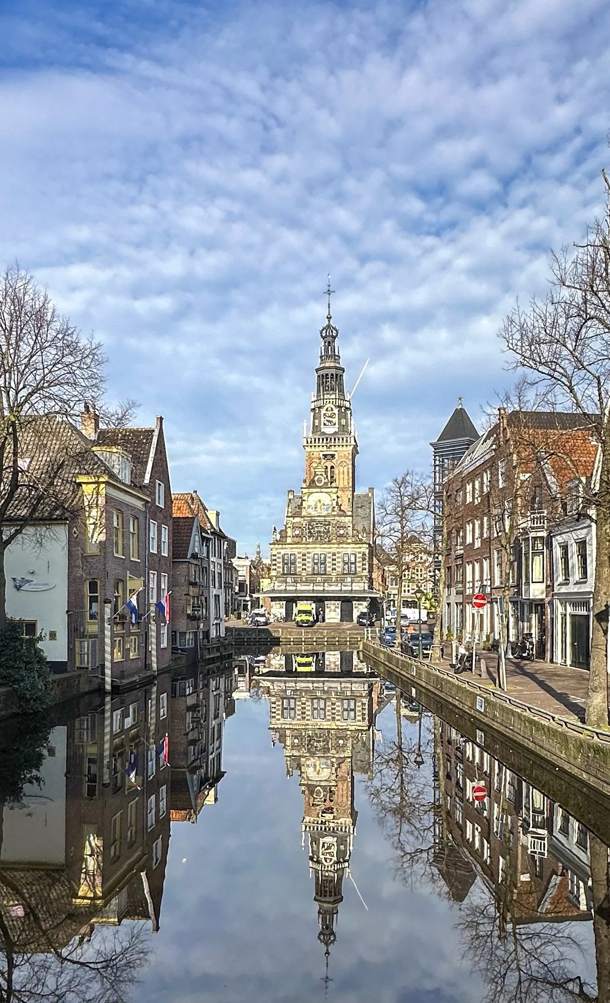 View of a historic clock tower with a reflection in the canal below, surrounded by traditional European buildings and a partly cloudy sky.