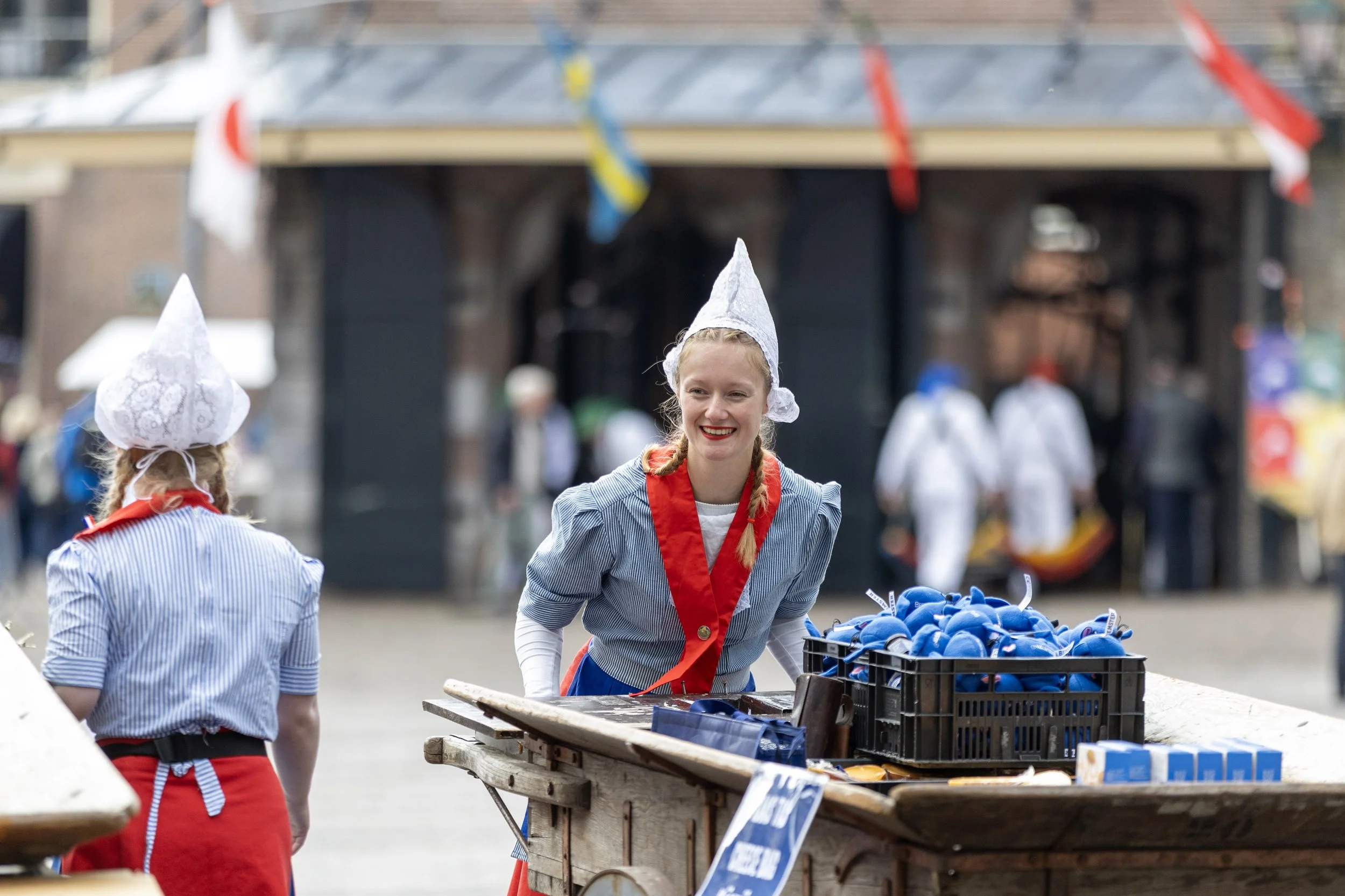 Women dressed in traditional Dutch clothing, including white lace caps and red scarves, selling blue slippers at an outdoor market during a festival or celebration.