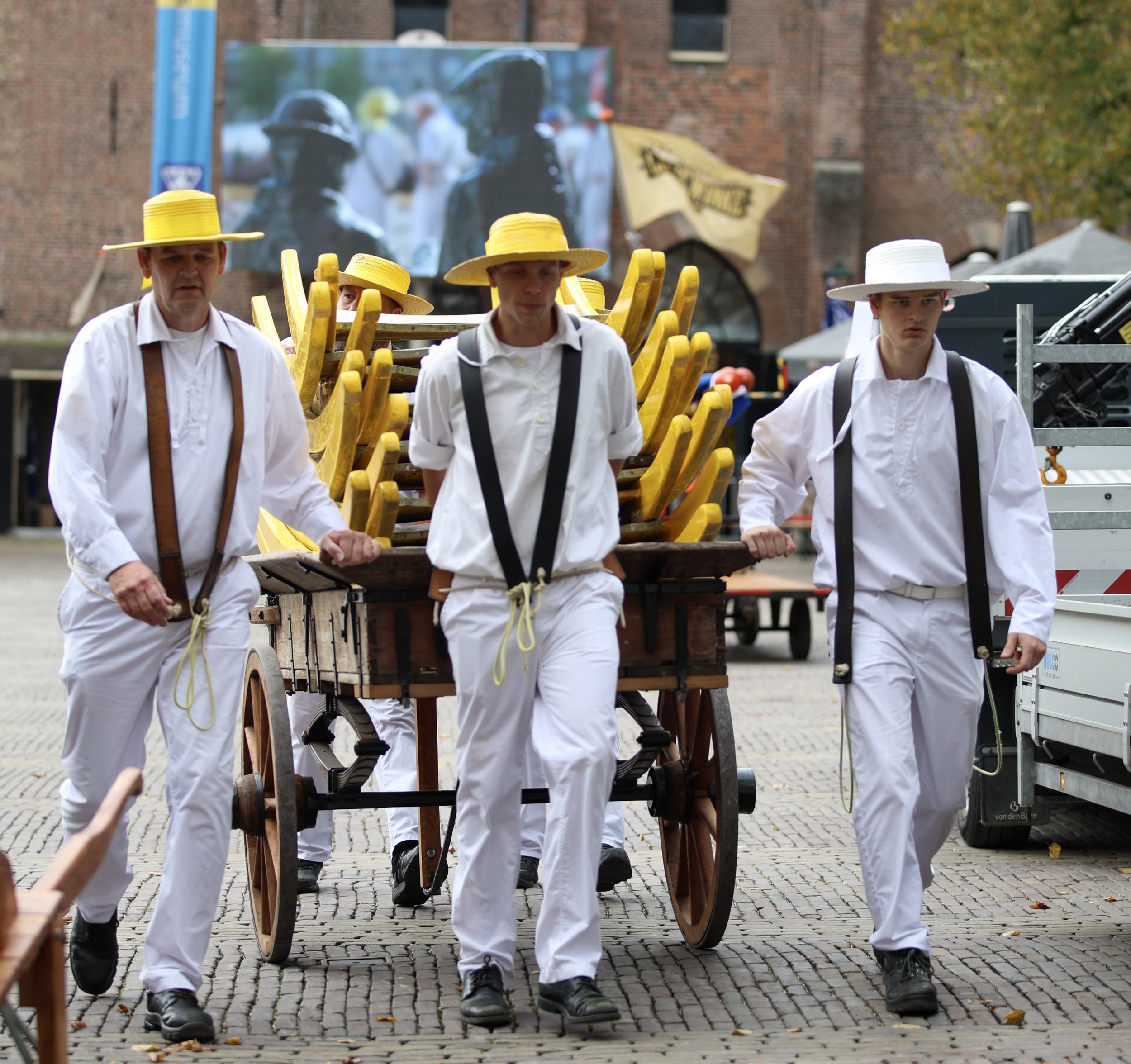 Group of people dressed in white with yellow and white hats, pulling a cart loaded with yellow wooden objects, walking on cobblestone street.
