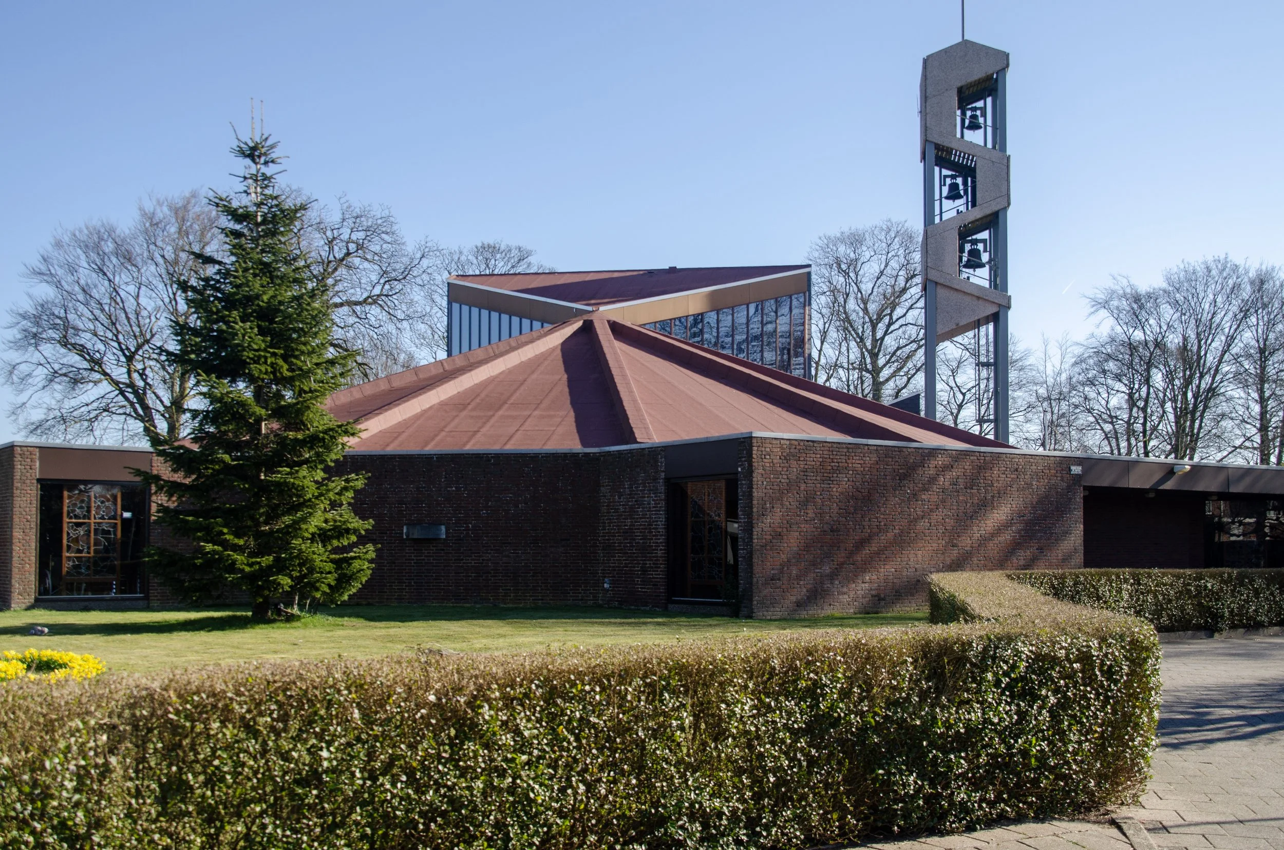 A church with a modern design, a brick facade, a tiered copper-colored roof, and a tall bell tower with multiple bells, surrounded by a well-maintained lawn and bushes, with leafless trees in the background under a clear blue sky.