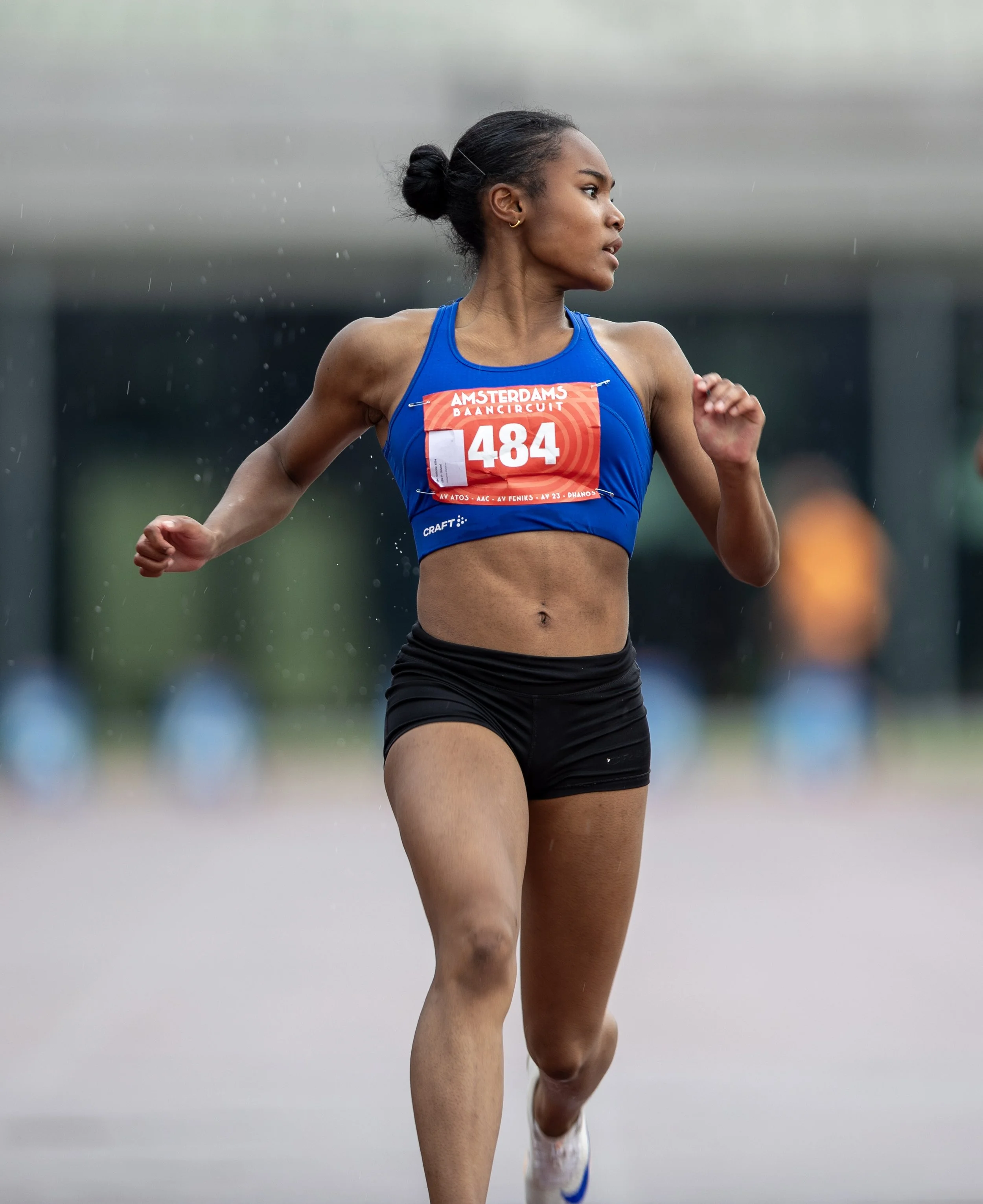 A female athlete running in a race, wearing a blue sports bra with a red race bib number 484 and black shorts, wet from rain.